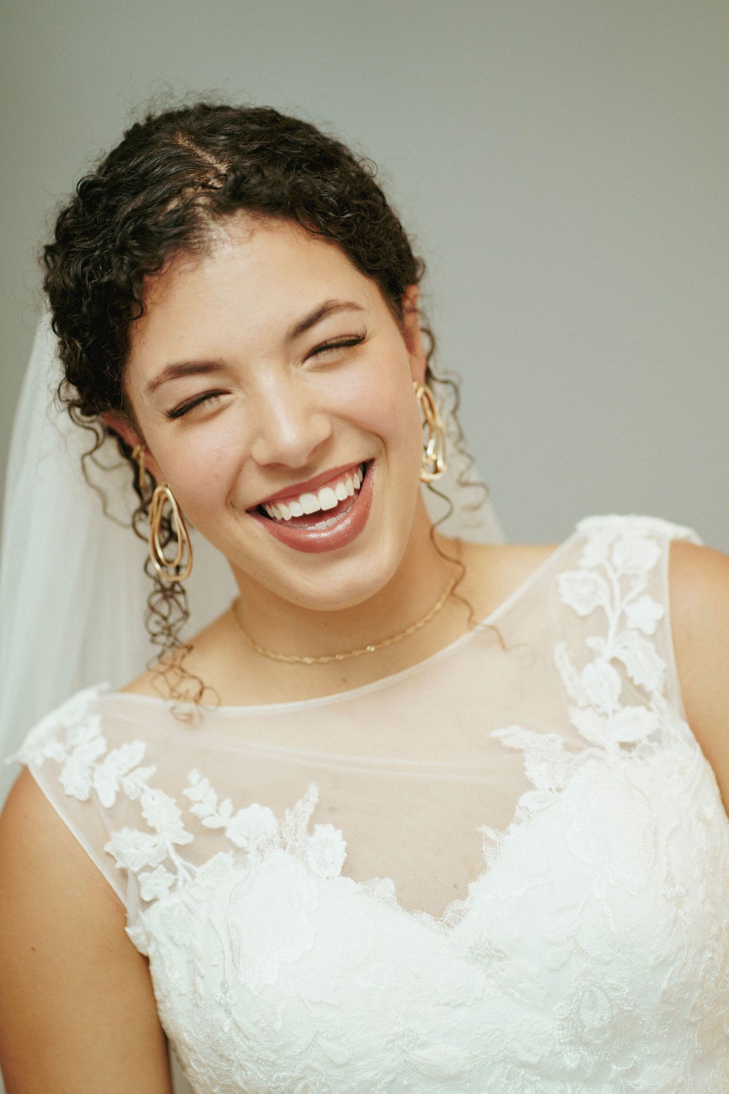 A joyful bride with curly hair and gold earrings laughs warmly while wearing a white lace wedding dress and veil.