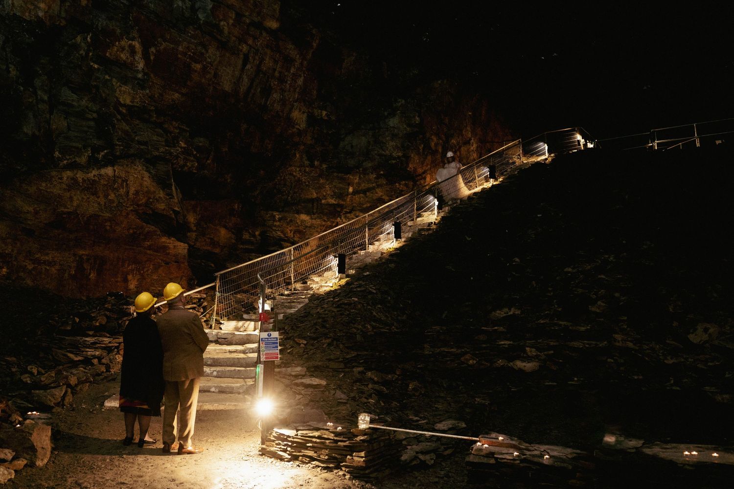 Two people in hard hats stand at the base of a lit staircase inside a dark volcanic cave.