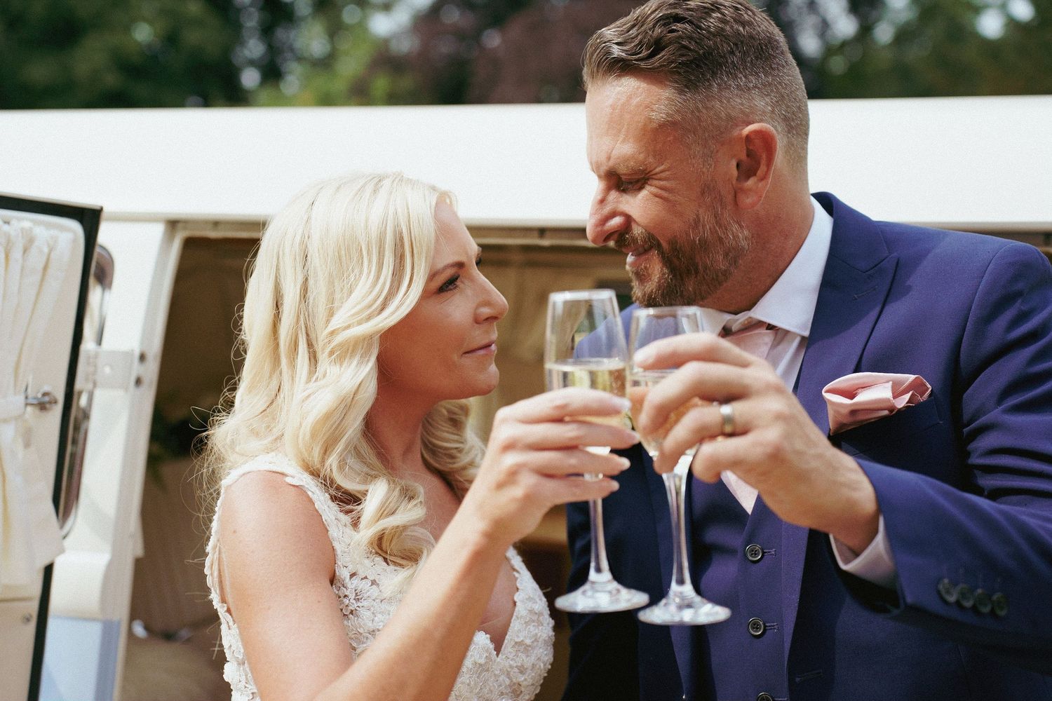 A blonde bride and a bearded groom in a blue suit smile at each other while toasting with champagne glasses.