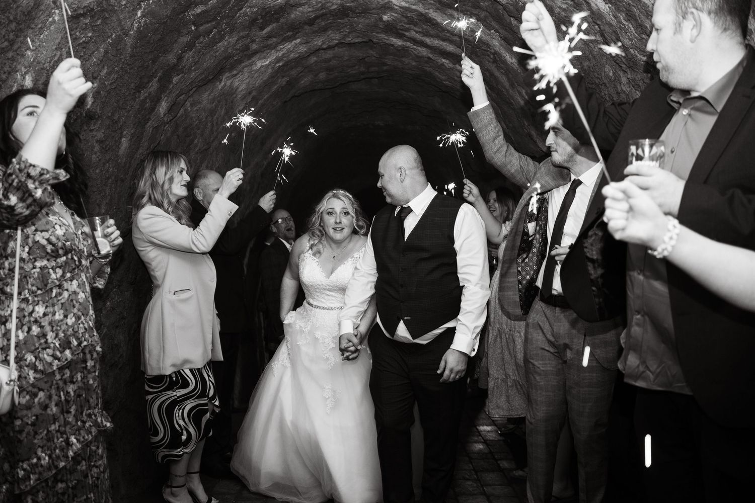 Bride and groom walk through a stone tunnel in black and white as wedding guests on both sides hold lit sparklers.