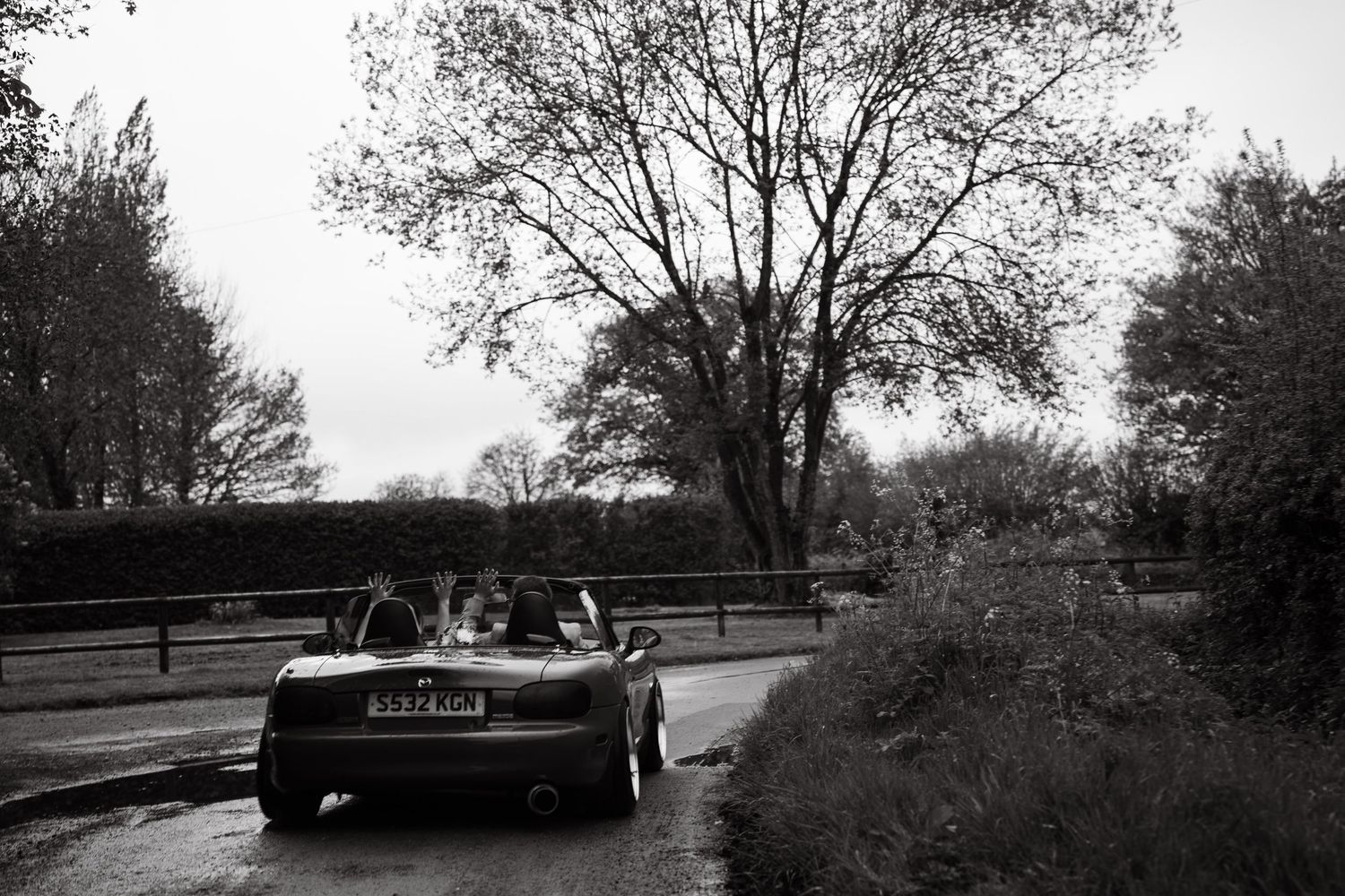 A black and white photo of a vintage convertible car carrying a wedding couple along a tree-lined country road.