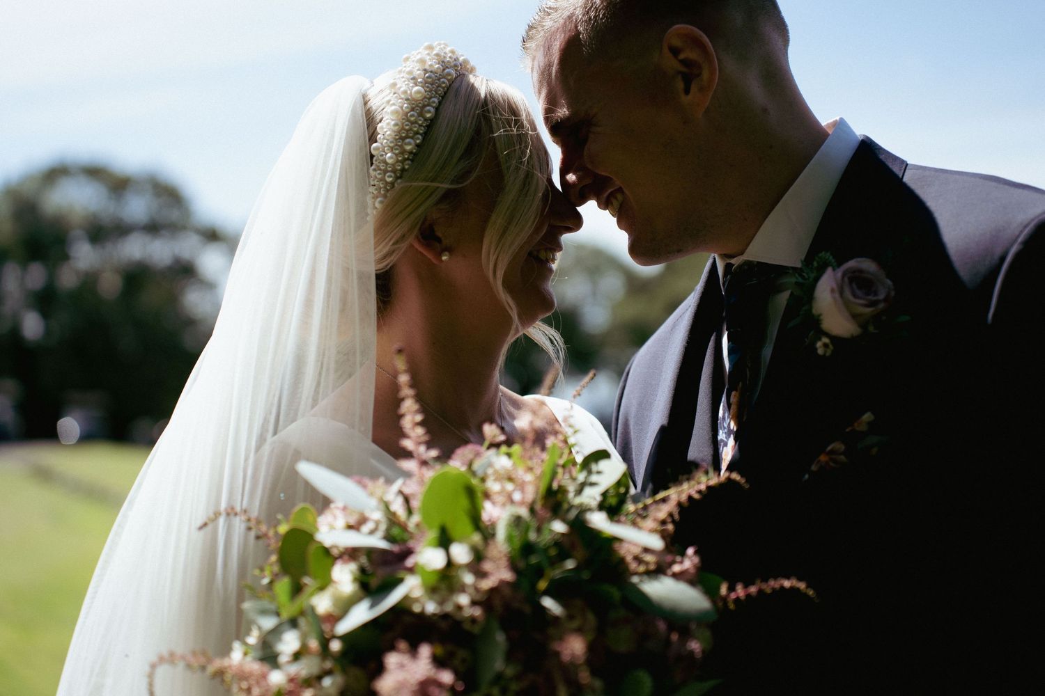 A bride and groom touch noses affectionately outdoors, holding a bouquet of pink and green flowers on their wedding day.