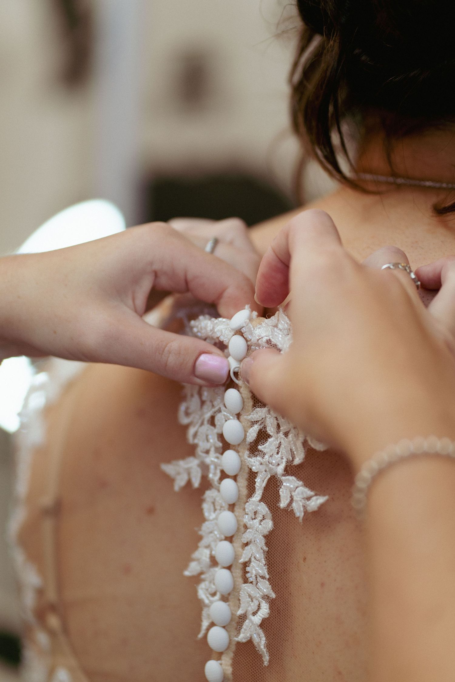 Two hands fasten the pearl buttons on the lace-trimmed back of a bride's wedding dress.