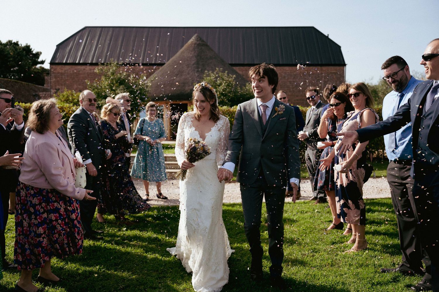 A bride in a lace gown and a groom in a gray suit walk hand in hand past cheering guests outside a rustic barn.