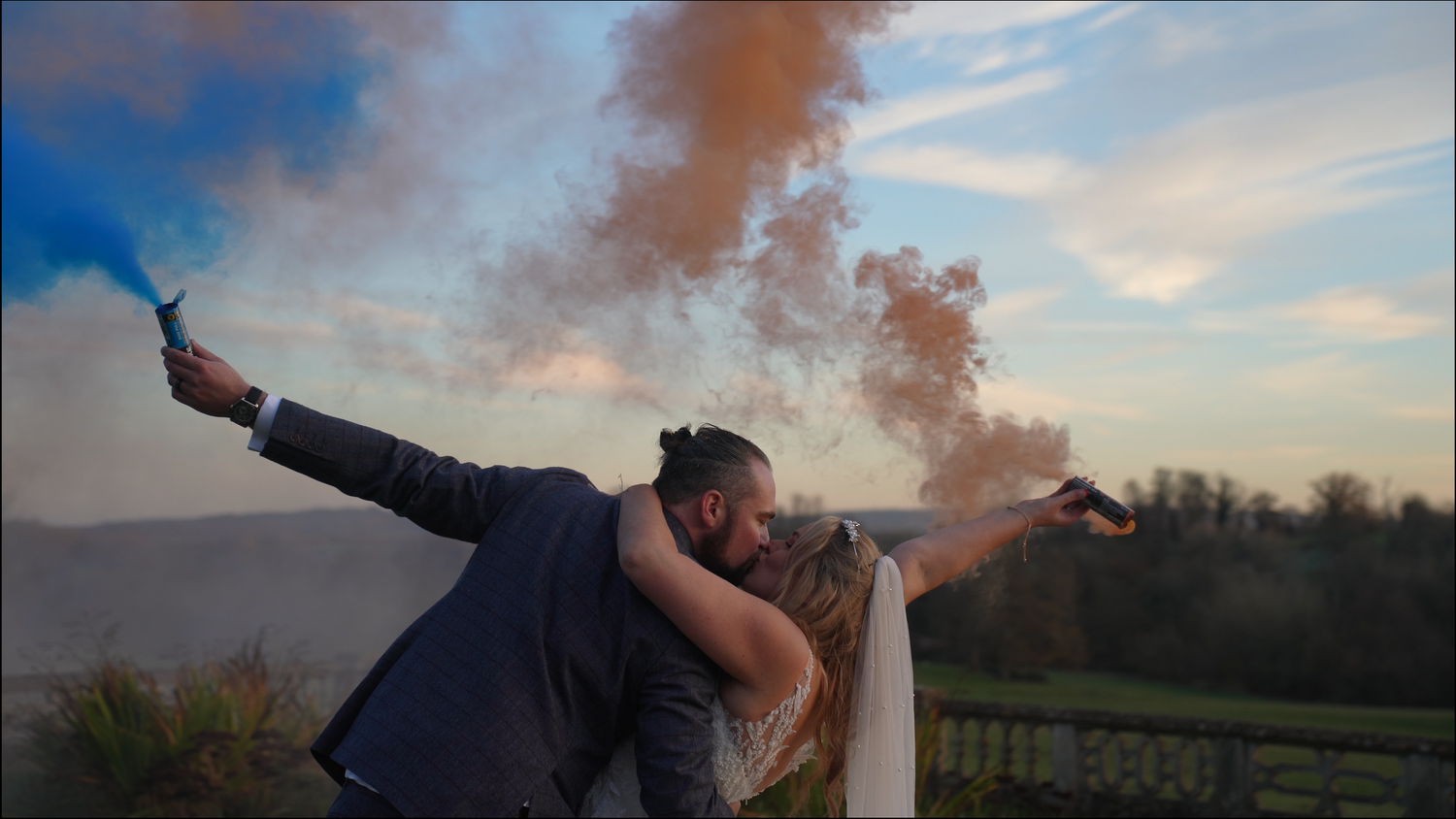 Bride and groom embrace at sunset while holding blue and orange smoke flares against a scenic hilltop landscape.