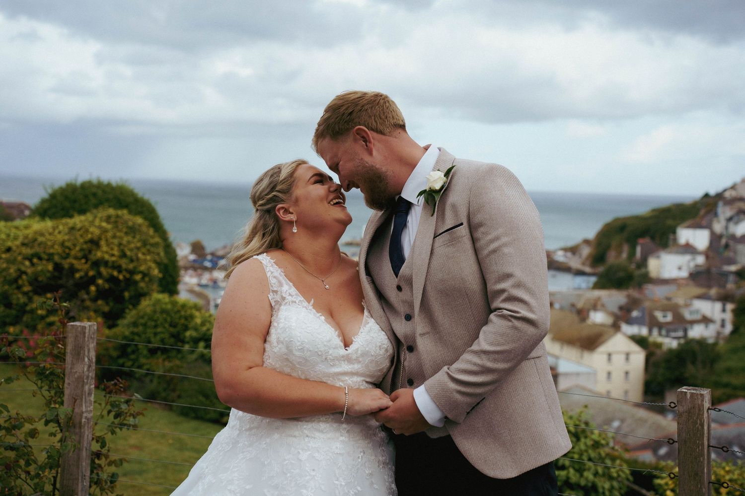 A bride and groom hold hands and smile at each other on a grassy hillside overlooking a coastal village and the sea.