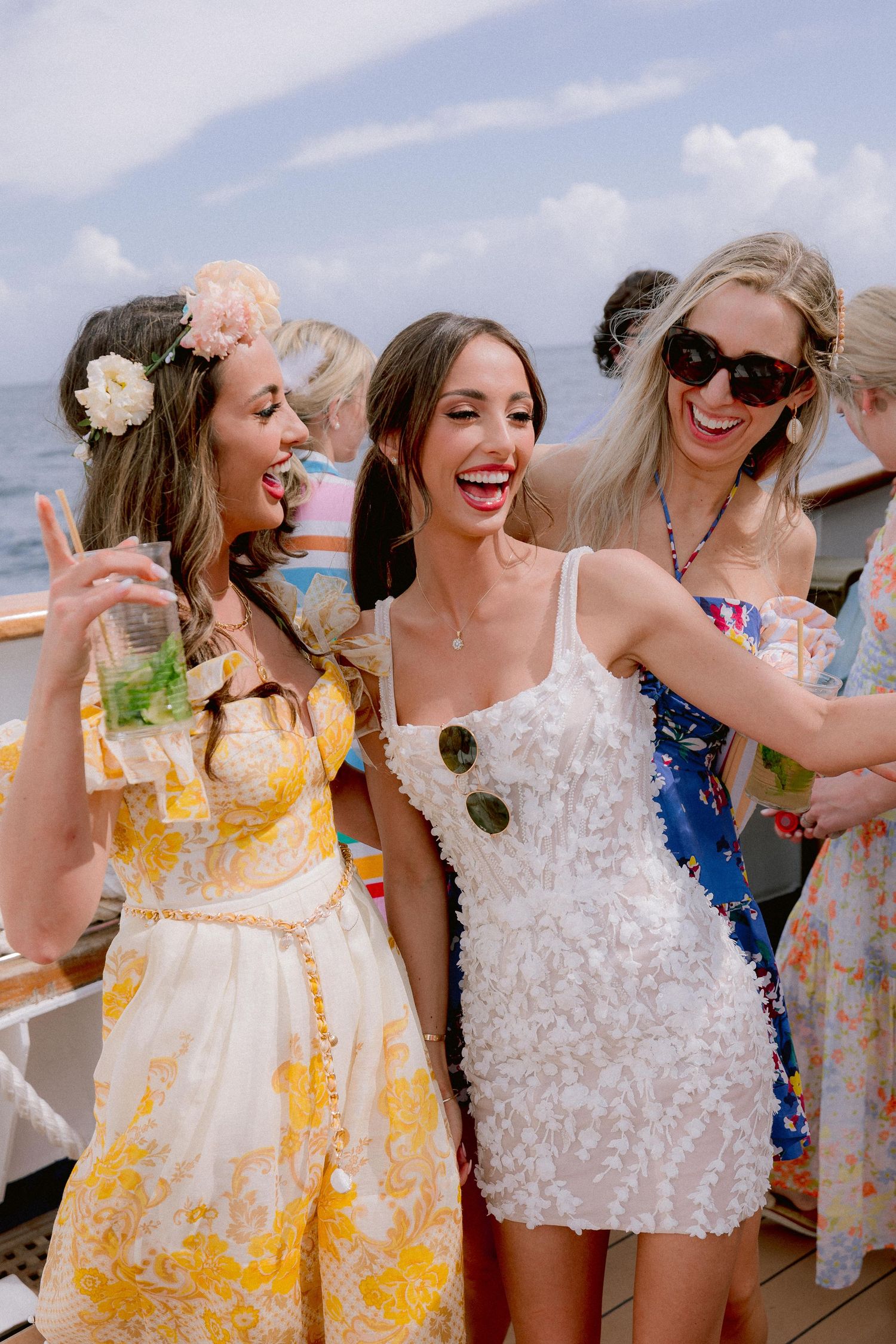 Three friends in summer dresses laugh and celebrate on a boat deck with tropical drinks and sunny skies.