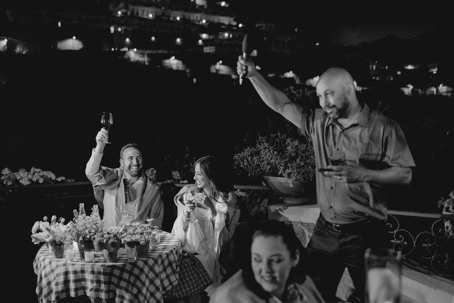 Black and white photo of a lively celebration scene at an outdoor party with people raising their arms in joyful gestures.