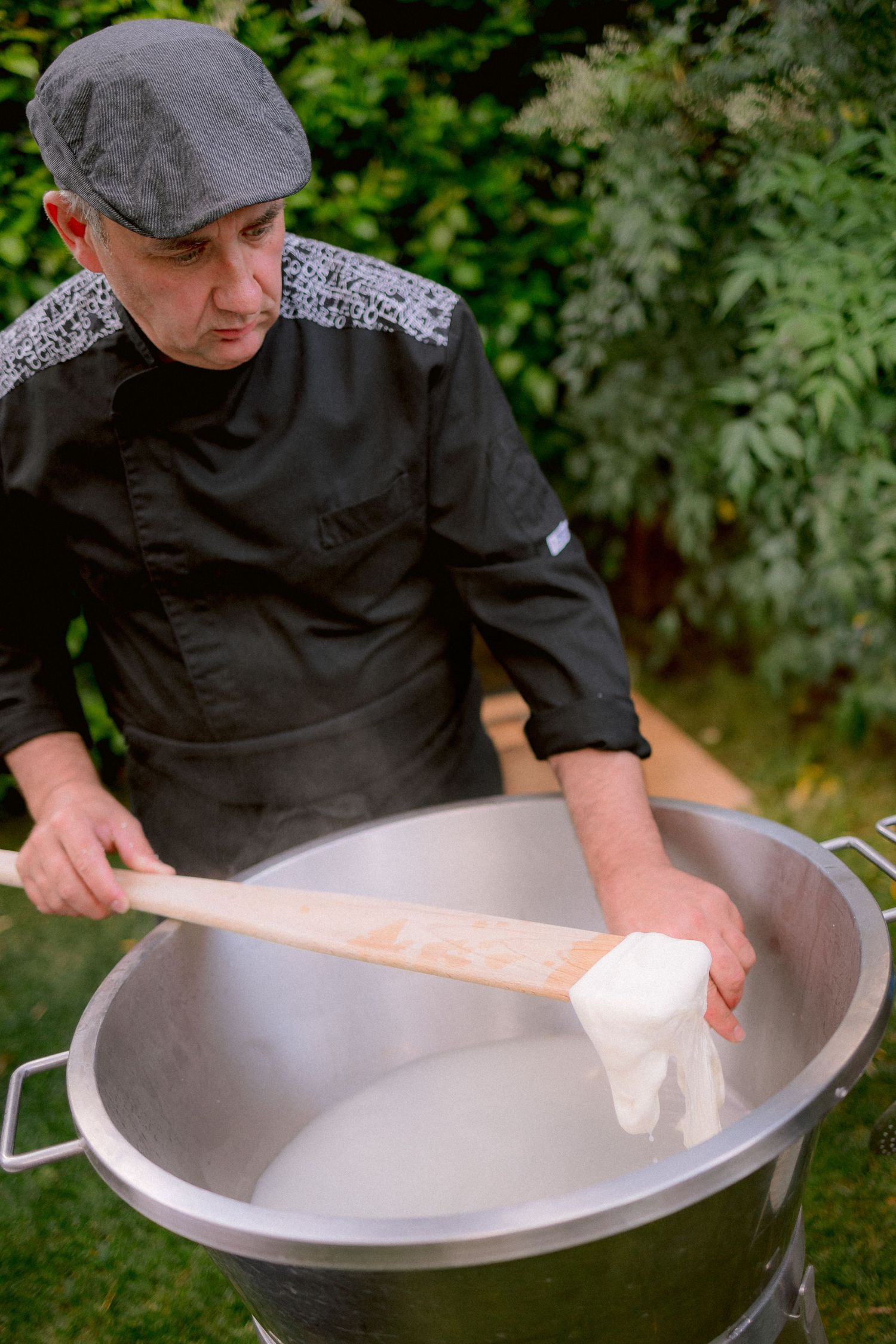 Chef in black uniform and grey cap prepares food in large metal cooking vessel outdoors in garden setting.