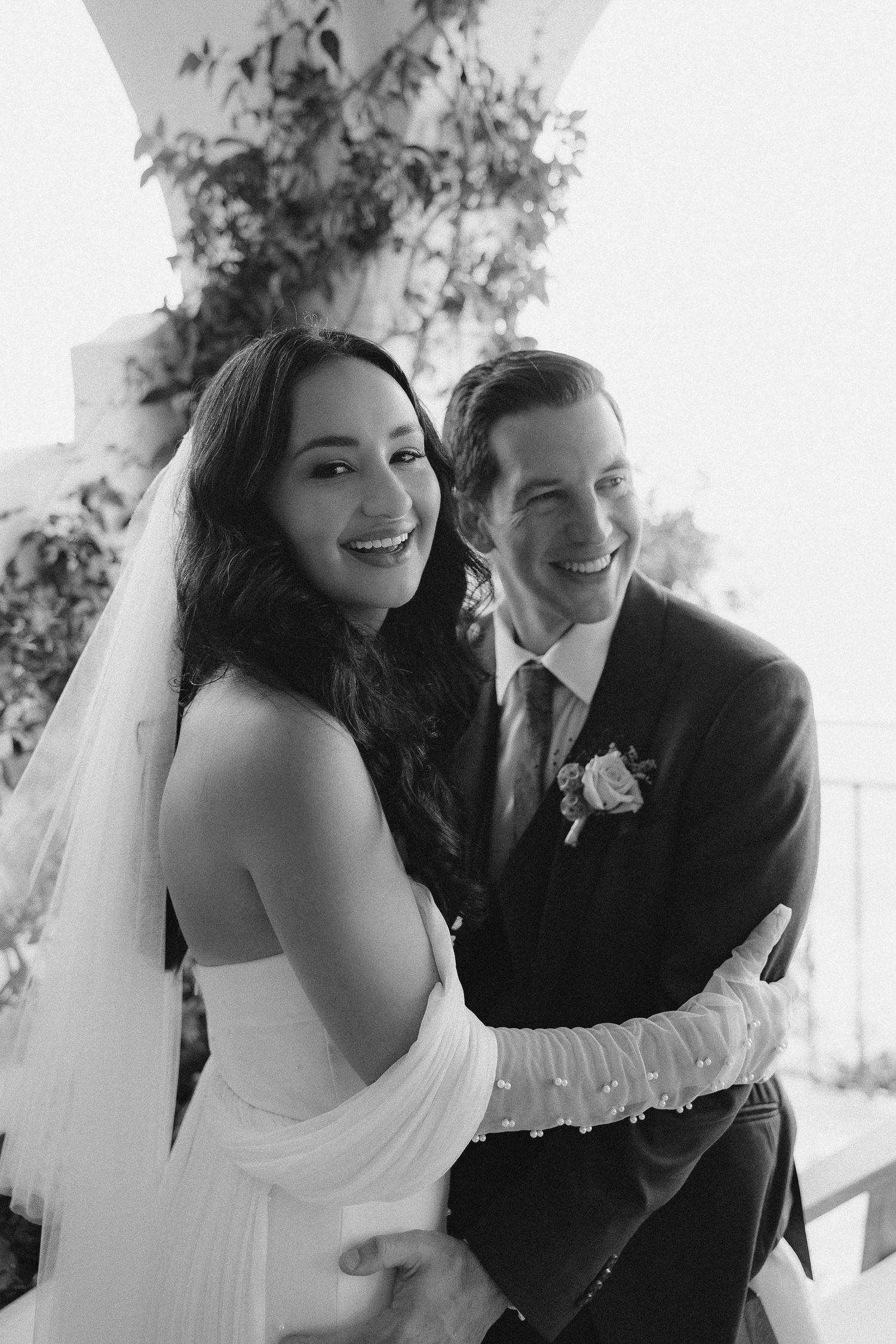 A black and white wedding photo of a bride in an off-shoulder dress and groom in a suit sharing a joyful moment.
