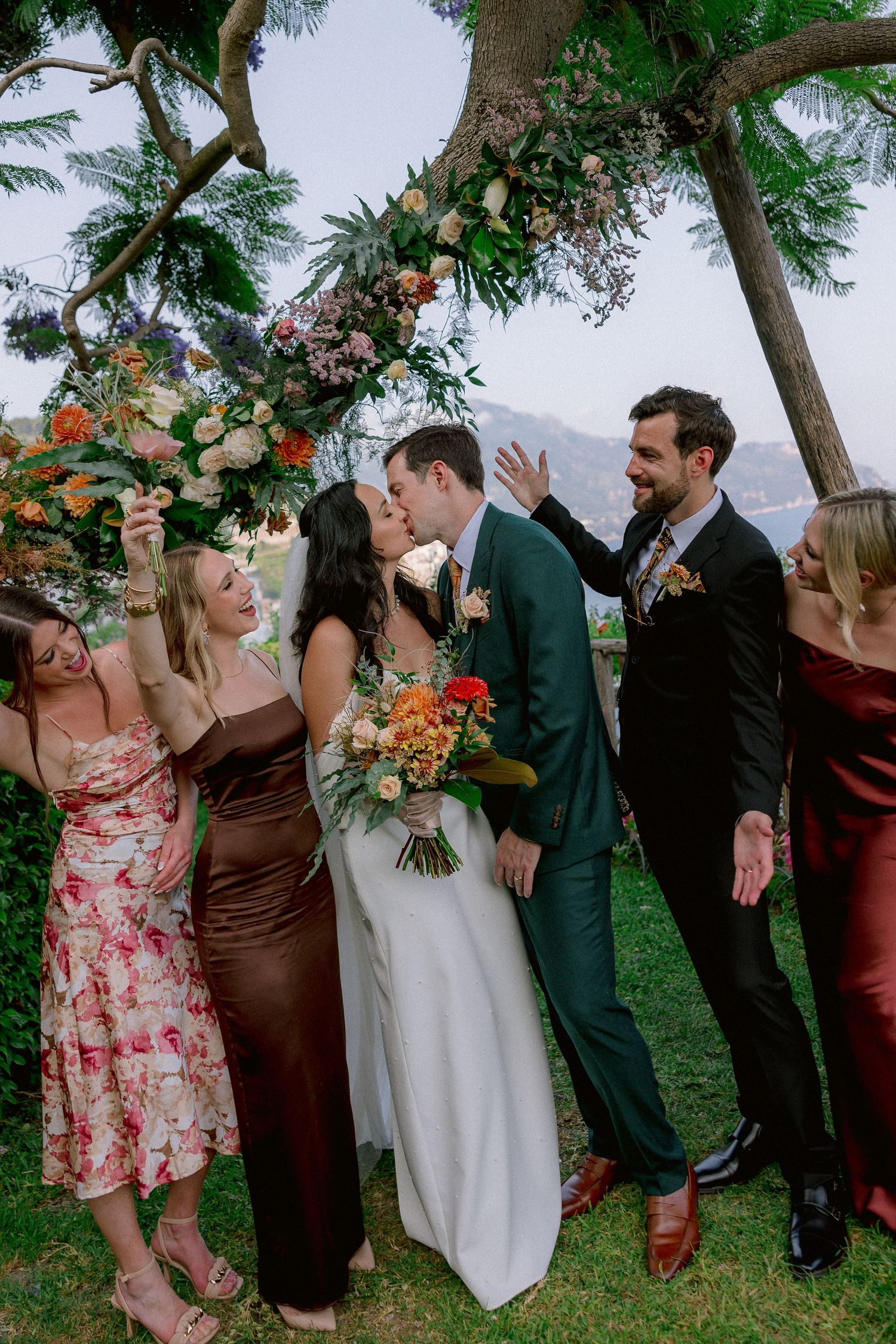 Wedding party poses under floral arch with autumn-toned decorations and greenery while celebrating outdoors.