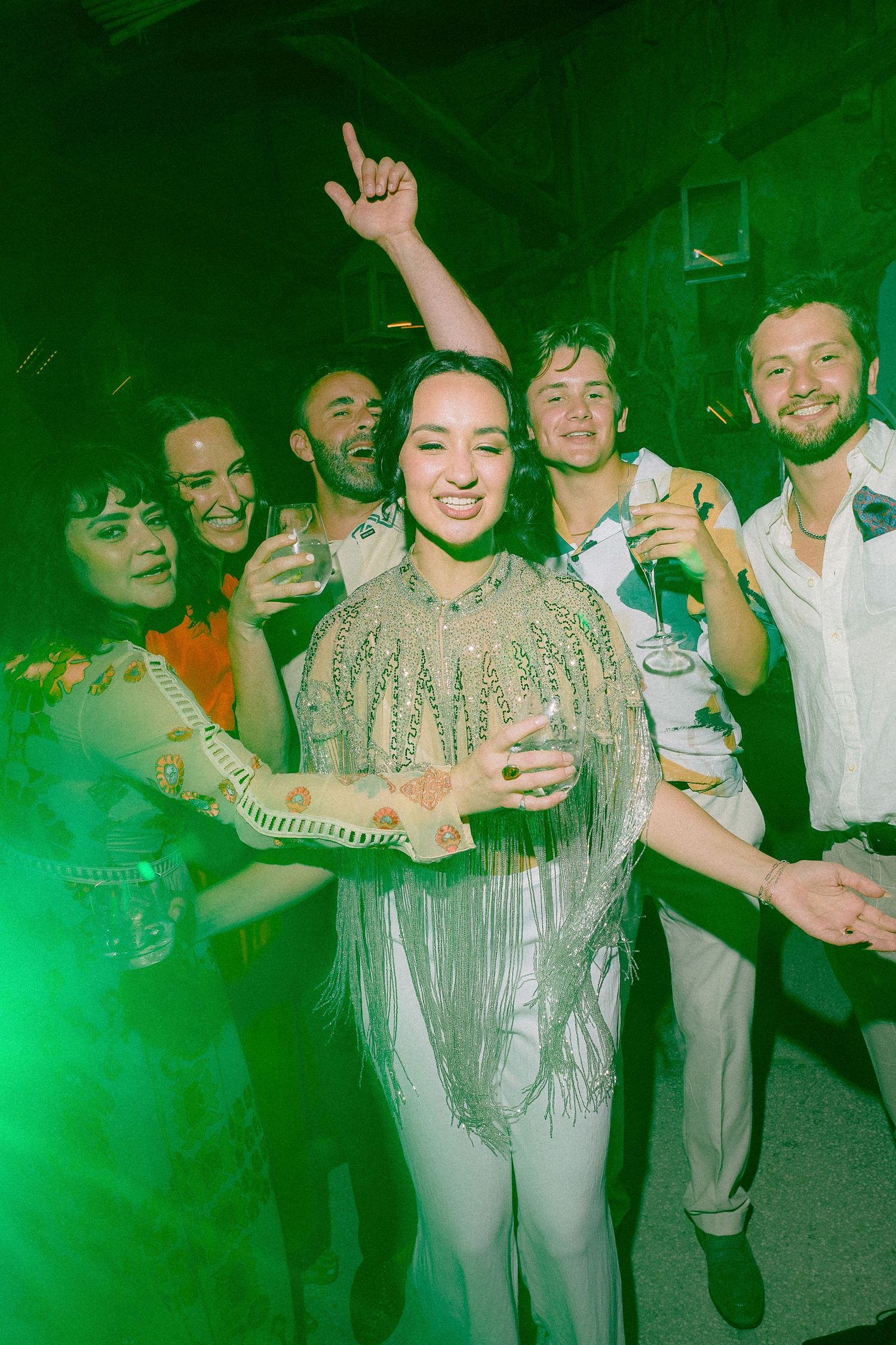 A group of people enjoying a party with green lighting as a person in a sparkly fringe outfit celebrates in the center.