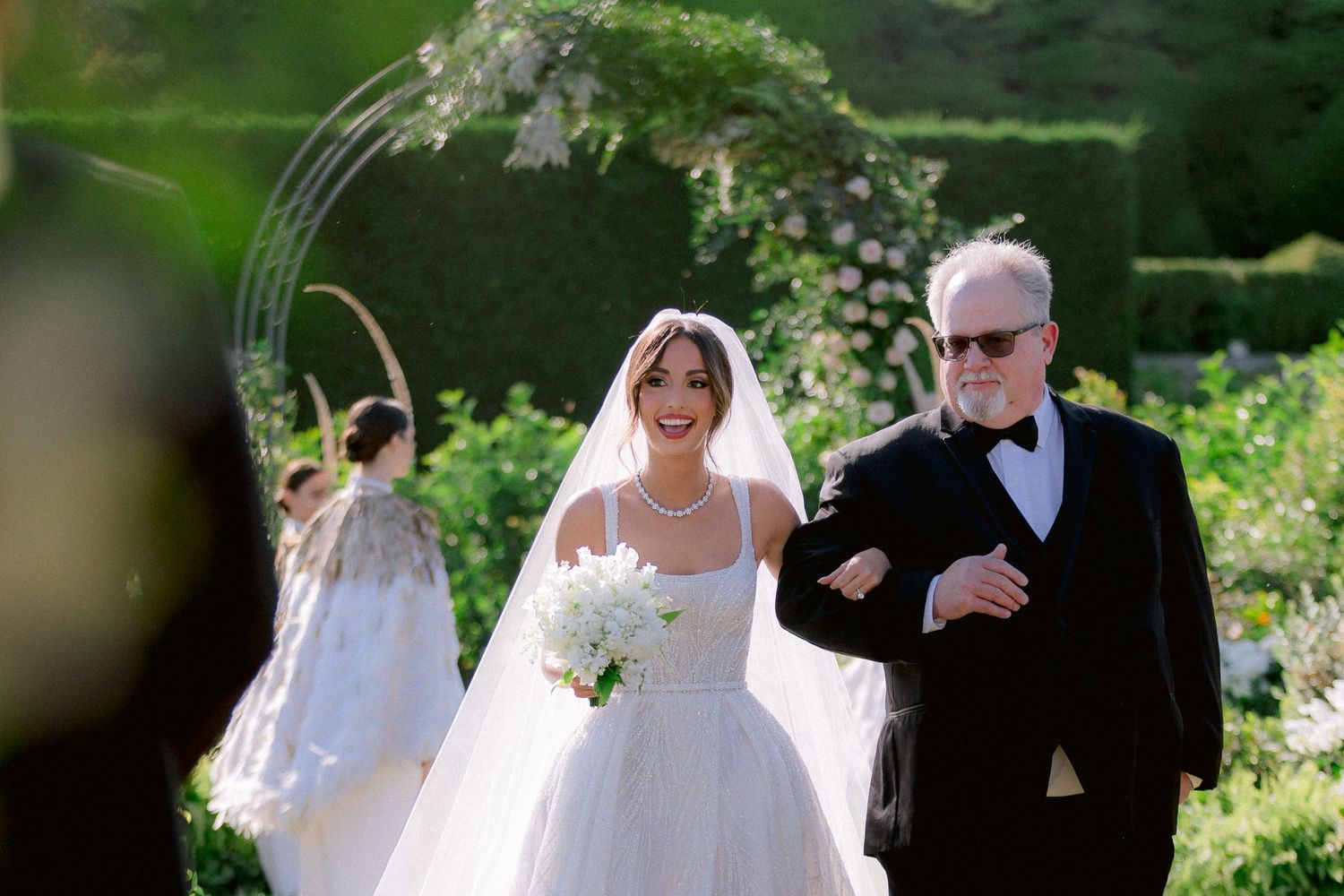 A traditional outdoor wedding ceremony with a bride in a white gown walking down a garden path under a floral archway.