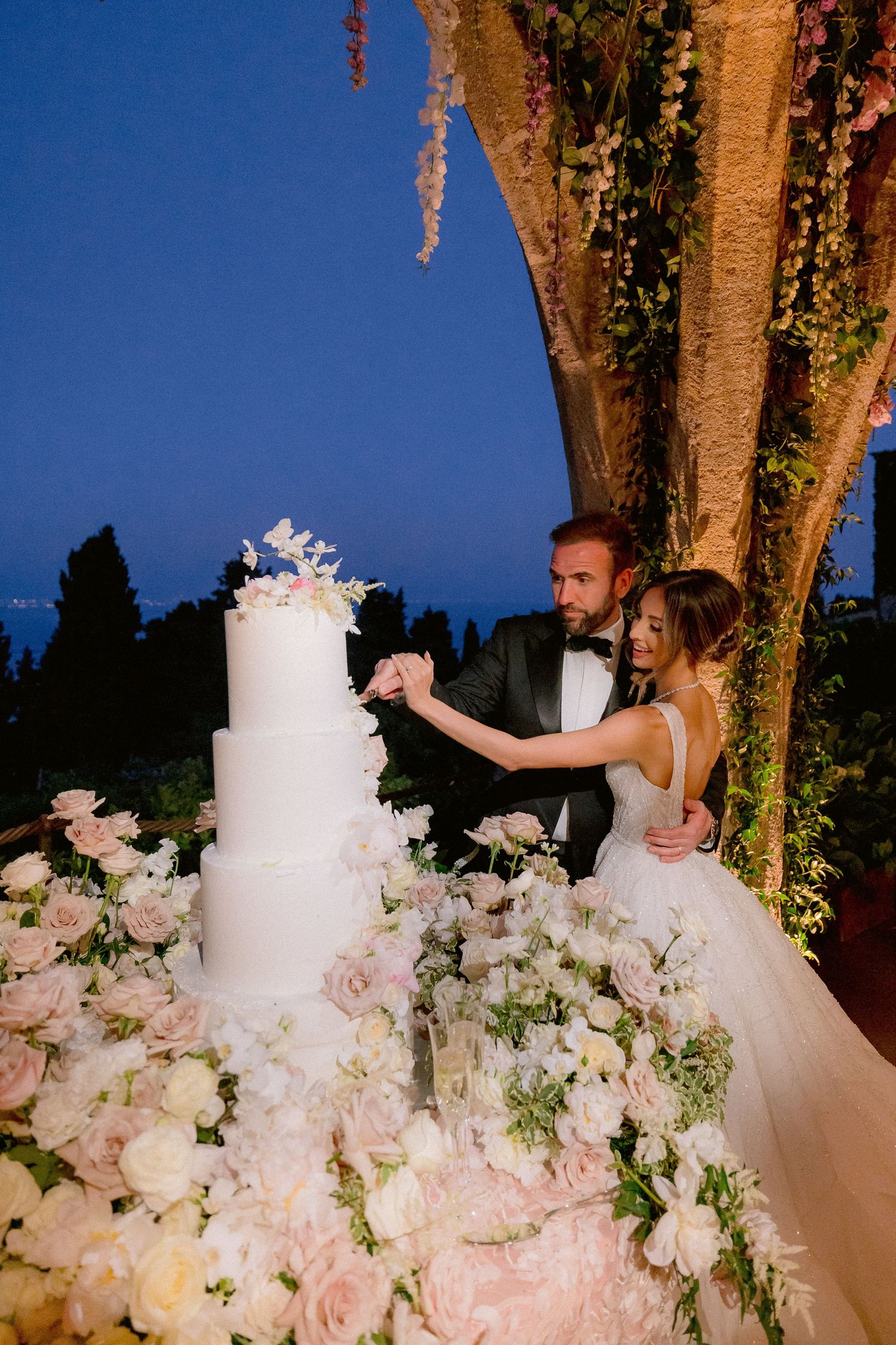 A traditional white wedding cake surrounded by blush pink and white flowers at an elegant evening outdoor reception.