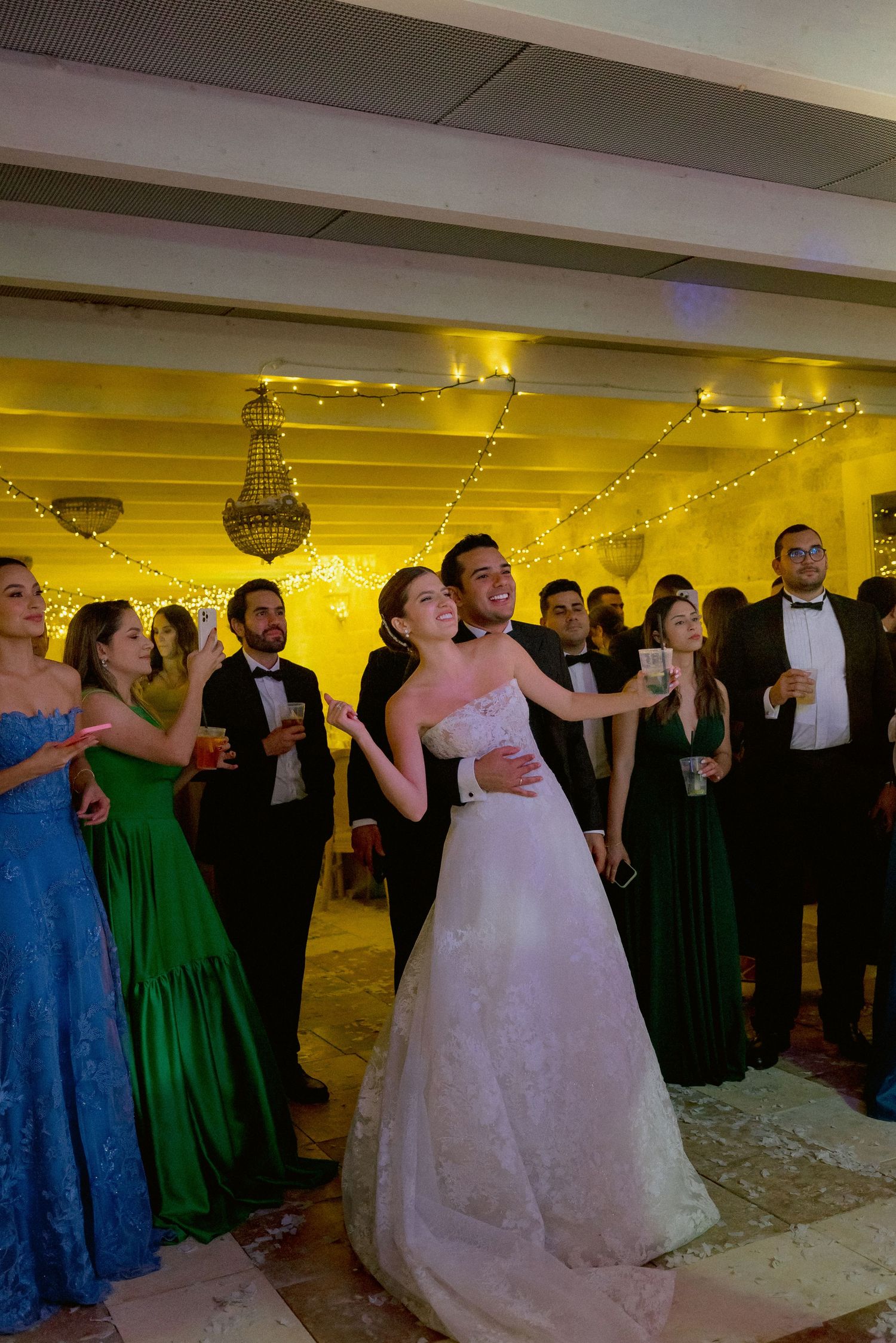 Wedding party guests celebrate on the dance floor under crystal chandeliers and string lights during the reception.