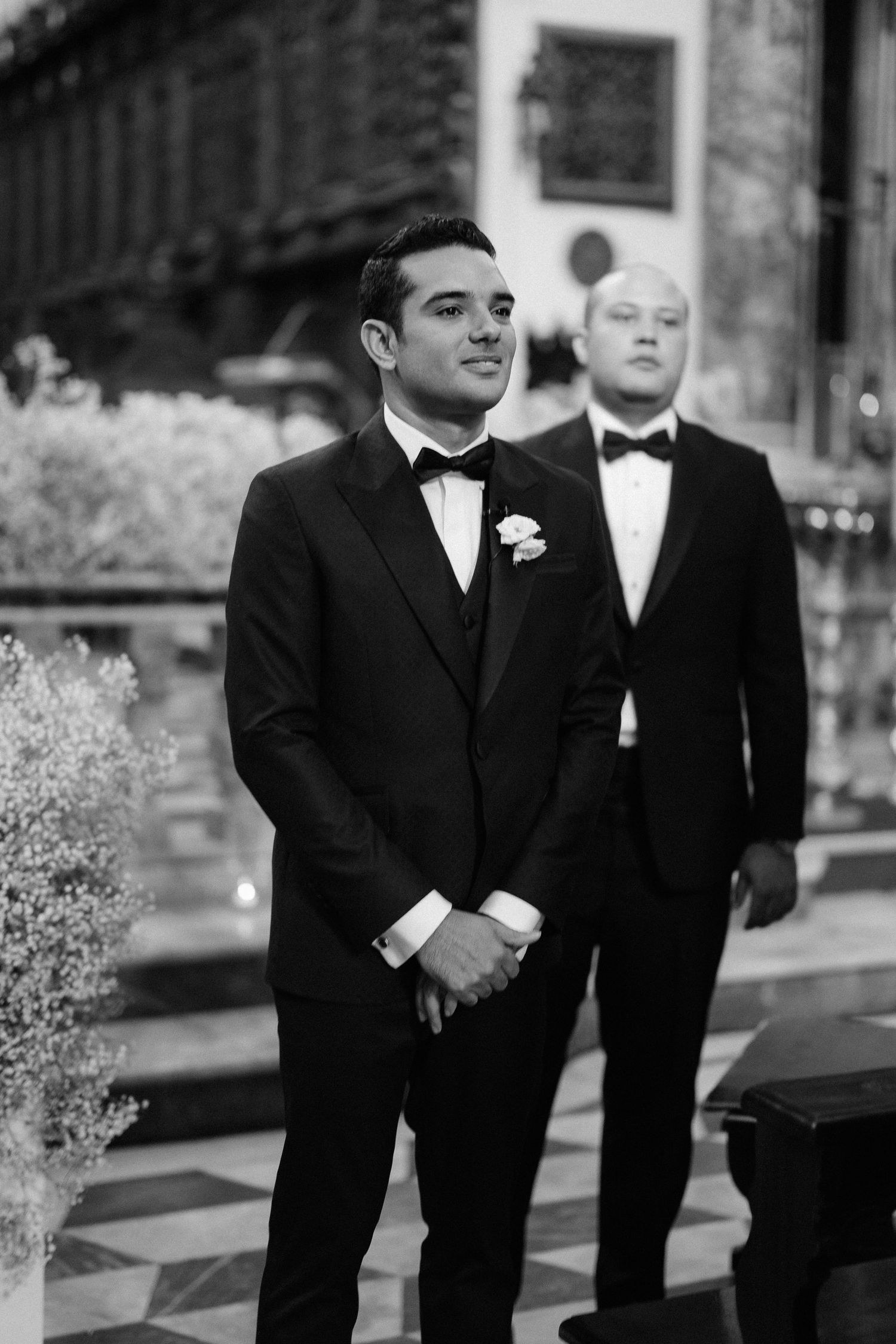 A black and white photograph of two men in tuxedos at a formal event, with a boutonniere on the groom.