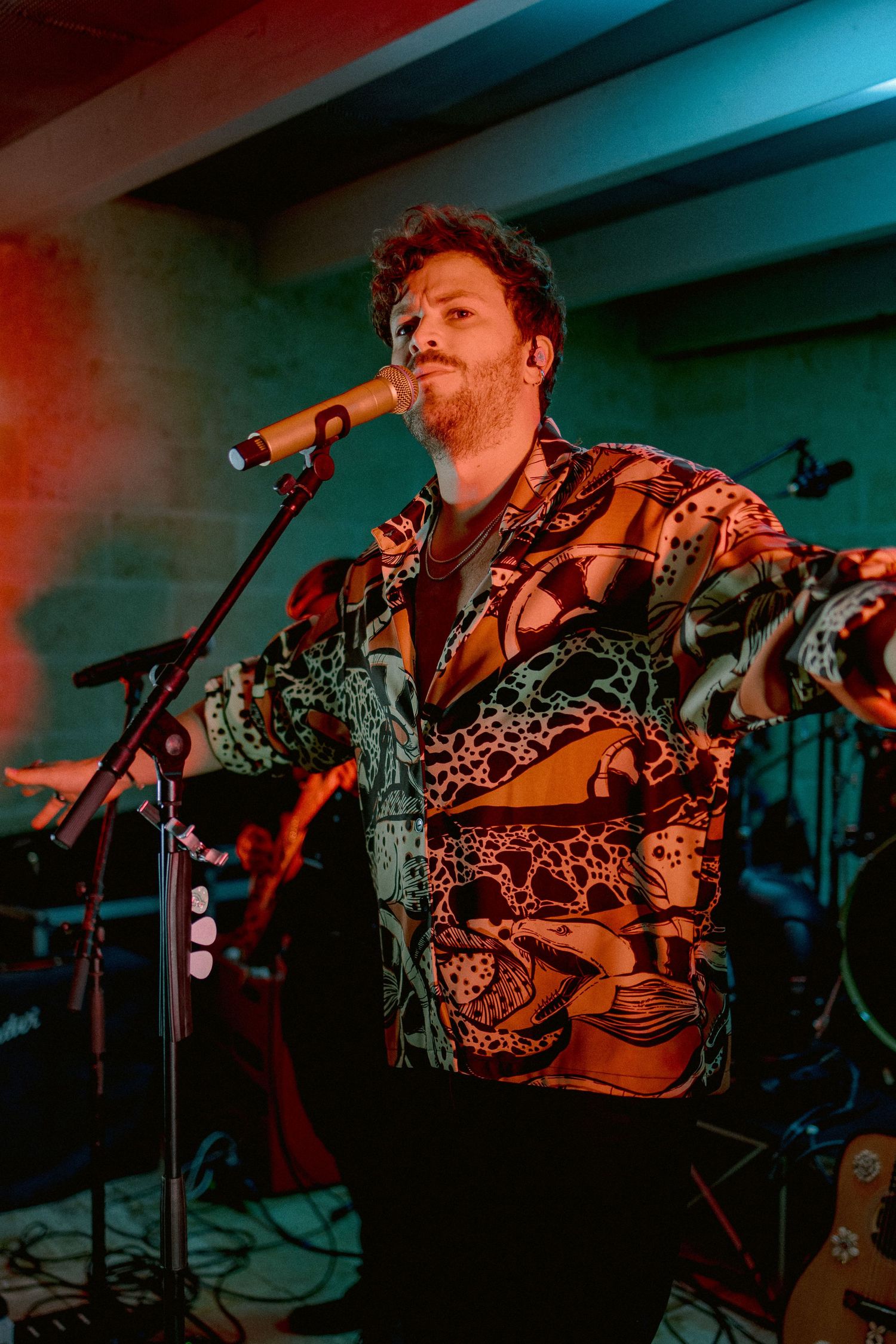 A performer in a patterned shirt sings into a microphone on stage with dramatic teal and orange lighting.