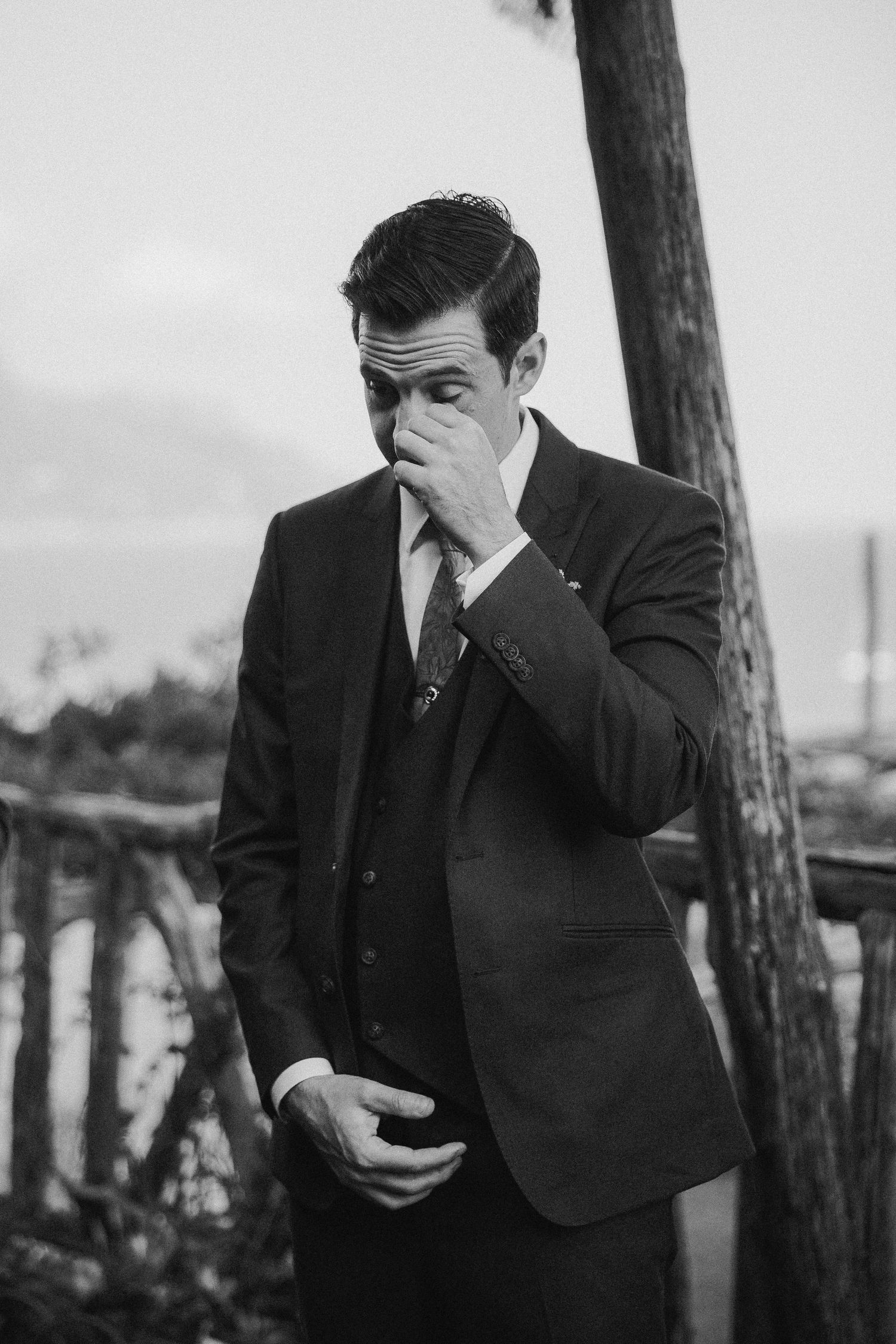 Black and white photo of someone in a dark suit covering their face with their hand while holding a phone.