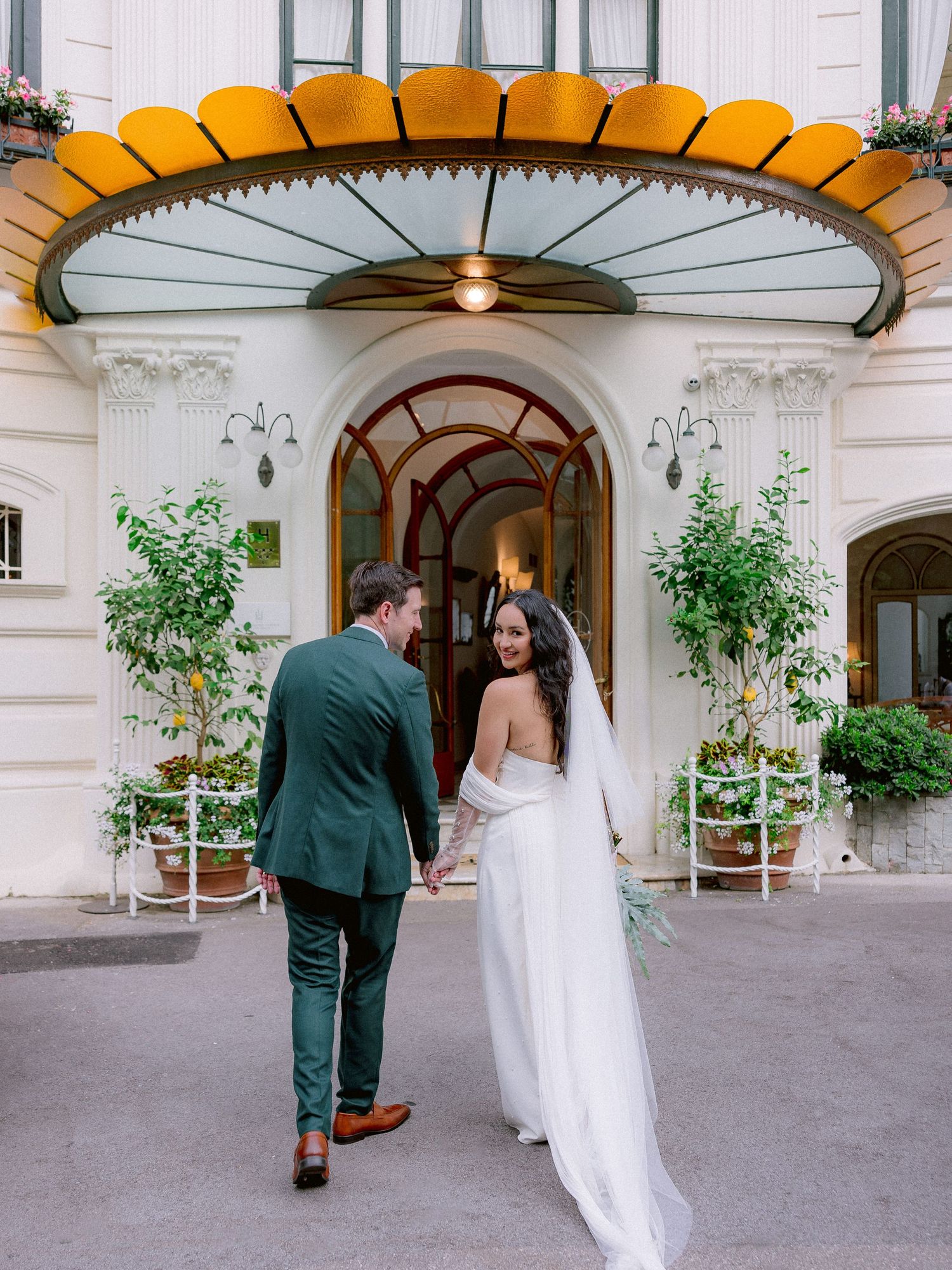 A wedding couple stands at the entrance of an elegant hotel with a distinctive orange-and-gold art deco awning.
