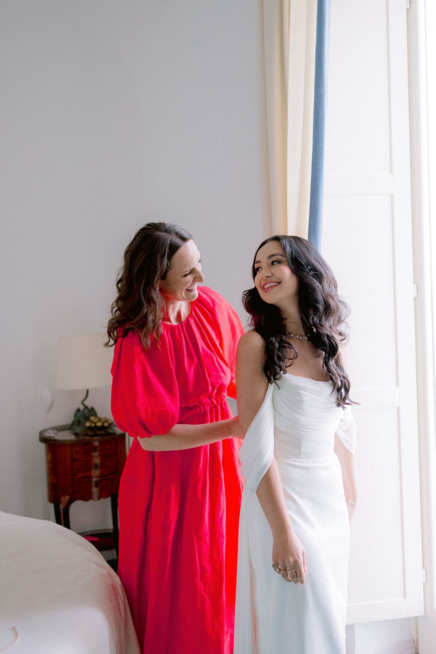 Two women sharing a joyful moment together, one wearing a bright red dress and one in an elegant white gown.