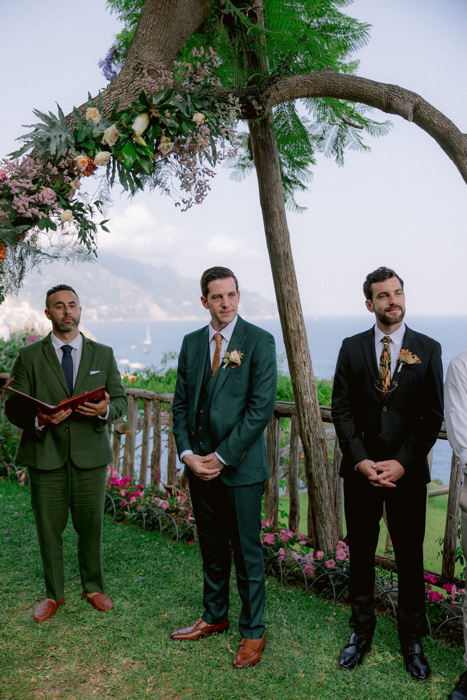 Three men in suits stand under a floral arch at an outdoor wedding ceremony with a scenic mountain backdrop.