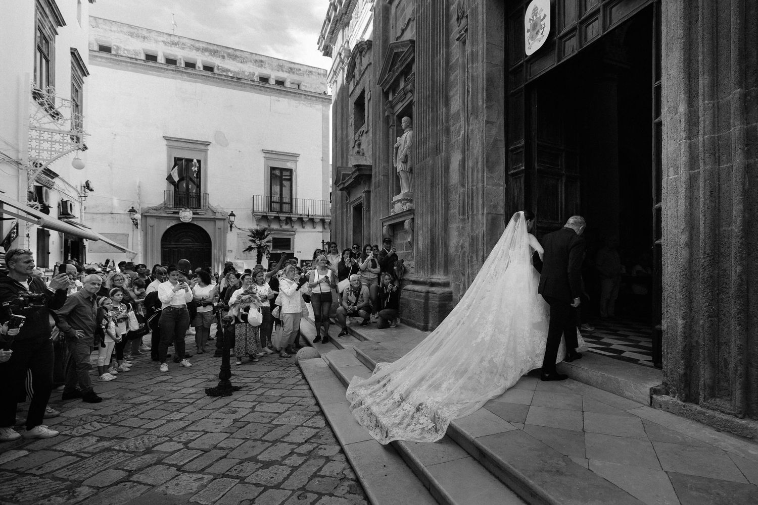 A black and white wedding photograph capturing a bride's train on church steps while guests look on from the plaza in Gallipoli, Puglia.