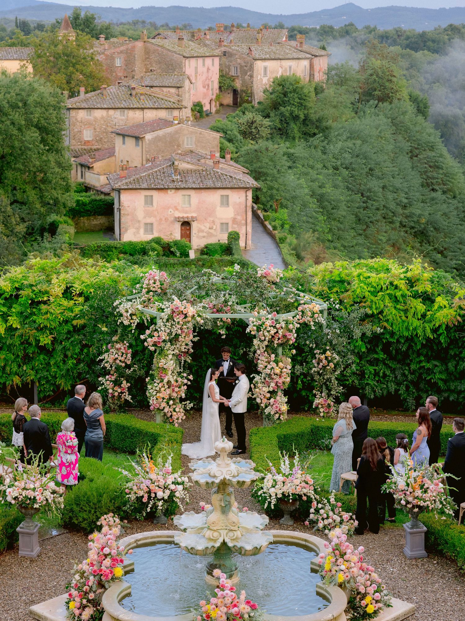 Romantic outdoor wedding ceremony in a Tuscan garden with floral arch, fountain, and historic Italian village backdrop.