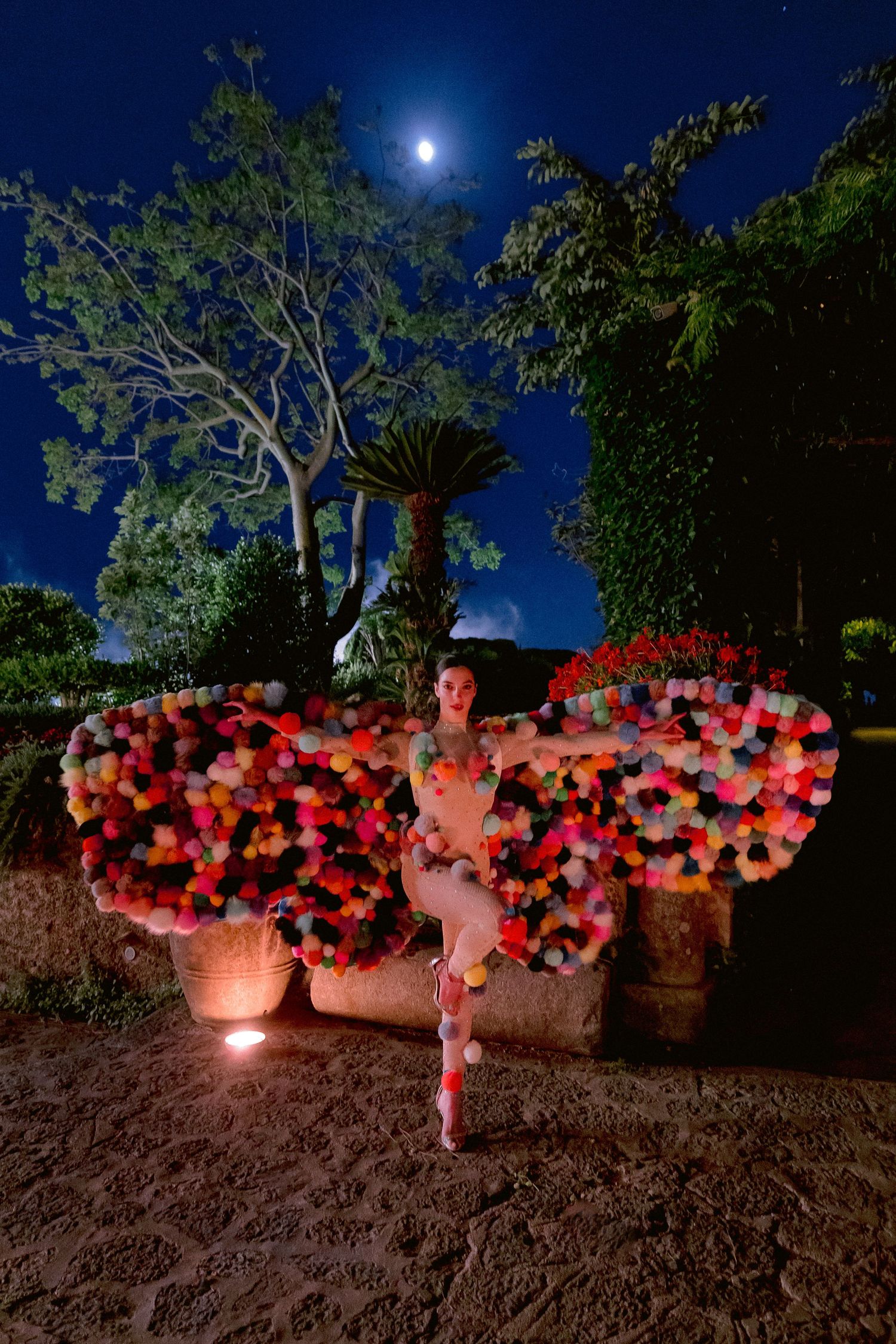A dreamy nighttime garden scene with illuminated colorful butterfly wings decoration and moonlight shining through trees.
