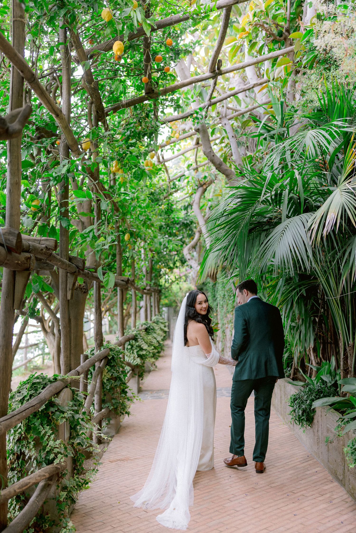 A couple walks along a romantic garden path under a trellis covered in greenery and yellow blooms at a wedding venue.
