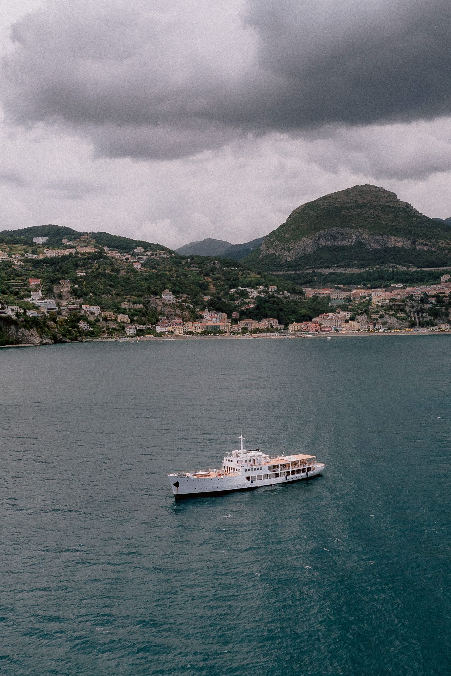 Ferry boat sailing through Mediterranean waters near coastal town with mountains and stormy clouds overhead.