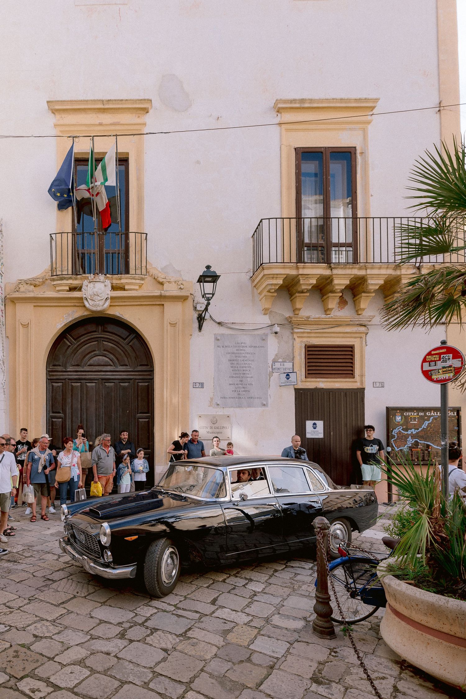 Vintage black luxury car parked in front of historic Italian building with traditional architecture and palm trees in Sicily.