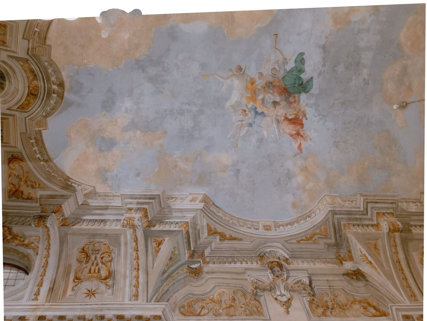 Ornate baroque church ceiling fresco featuring angels in flight amid clouds, with gilded architectural details and decorative moldings.