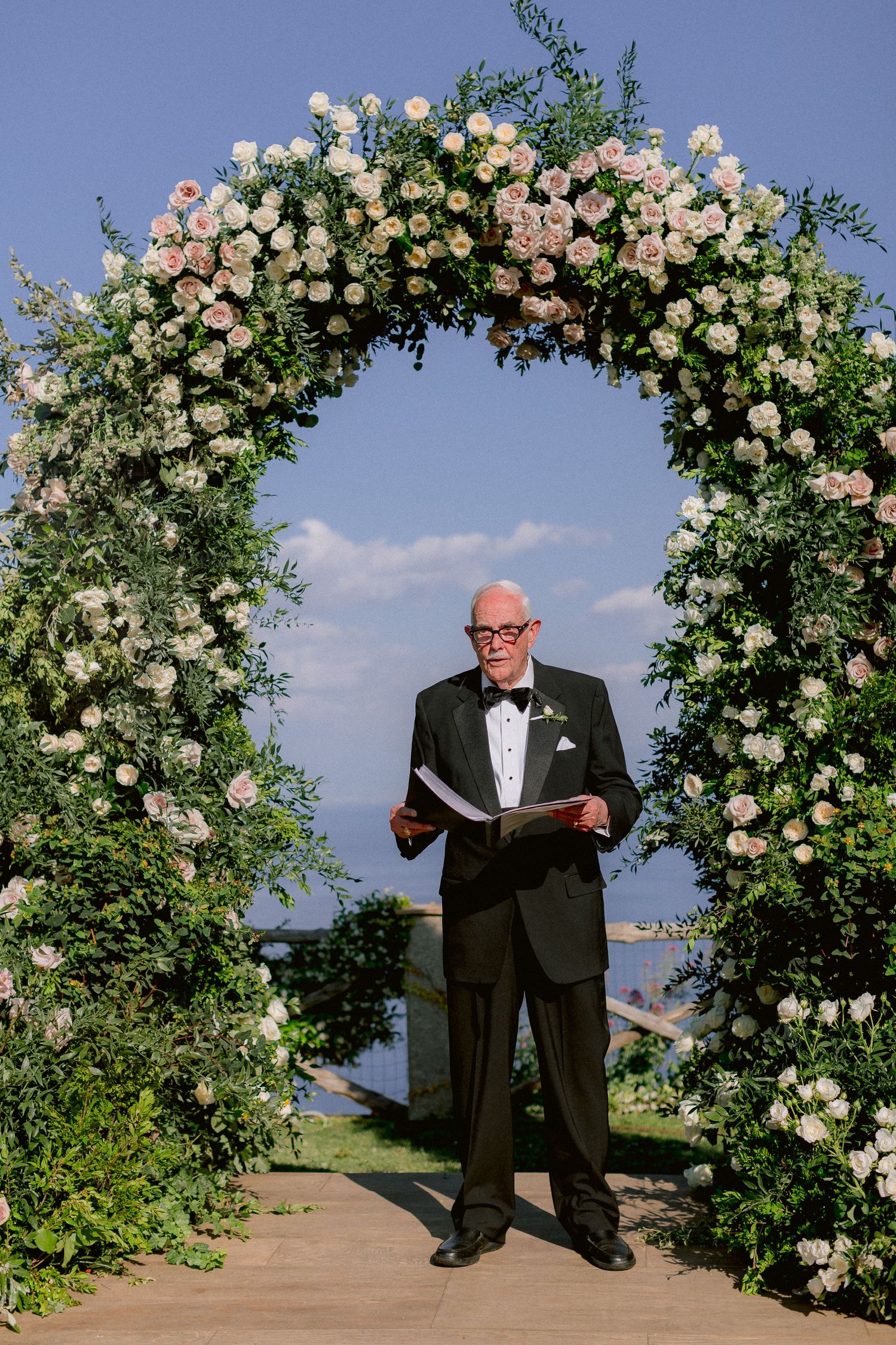 A floral wedding arch with white and pink roses frames a person in a black tuxedo holding a book outdoors.