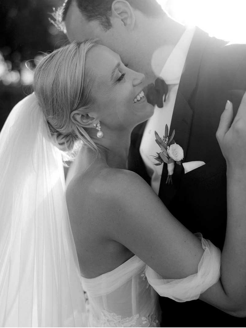 Black and white wedding photo of smiling bride in veil embracing groom in tuxedo with boutonniere.