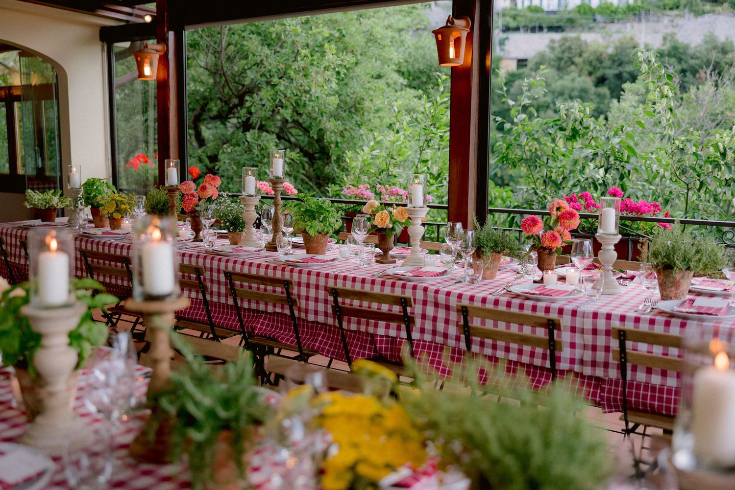 Long banquet table with red checkered tablecloths, candles and floral centerpieces on outdoor covered patio overlooking greenery.