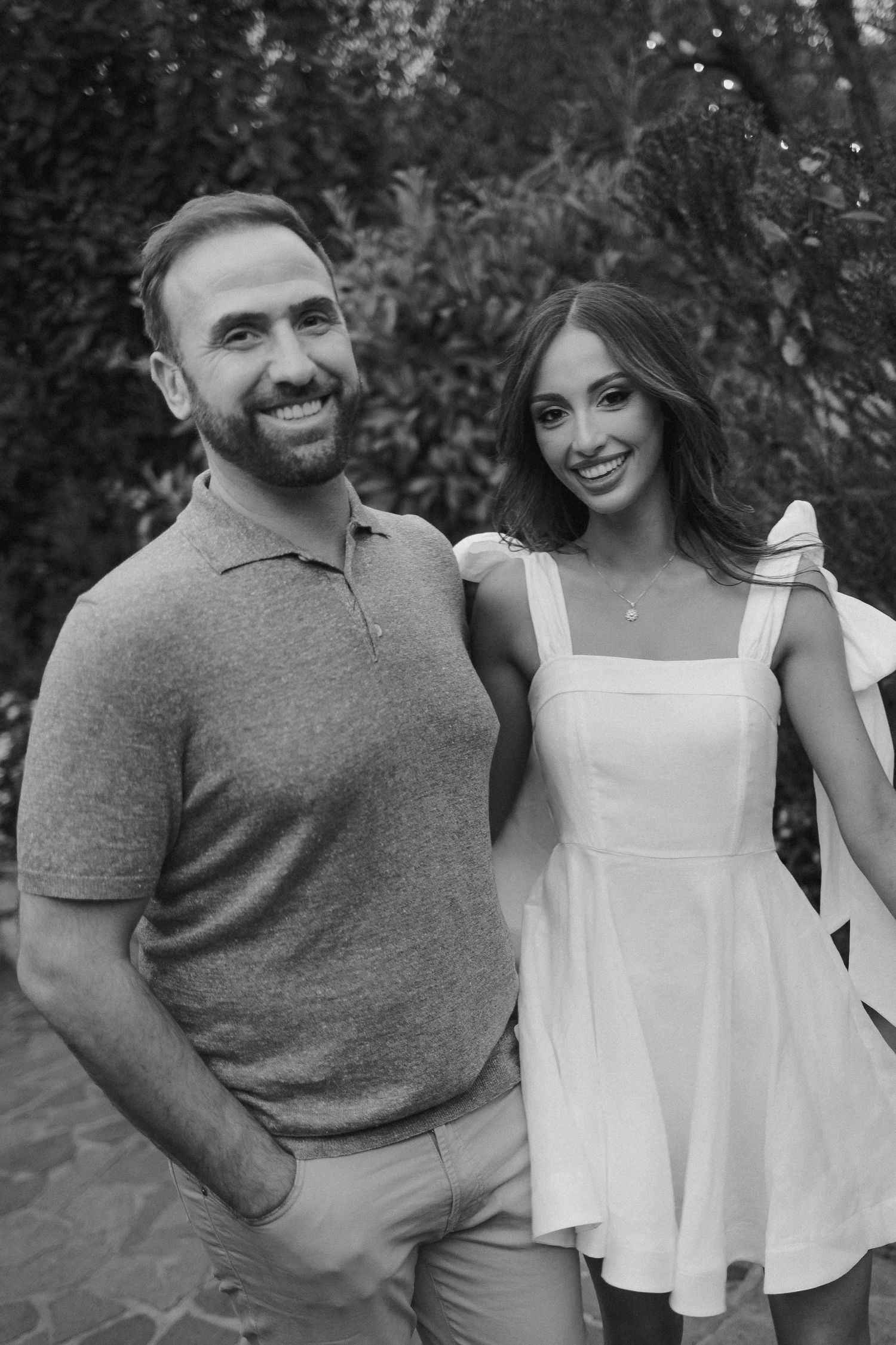 A smiling couple poses outdoors in black and white, with him wearing a polo shirt and her in a white summer dress.