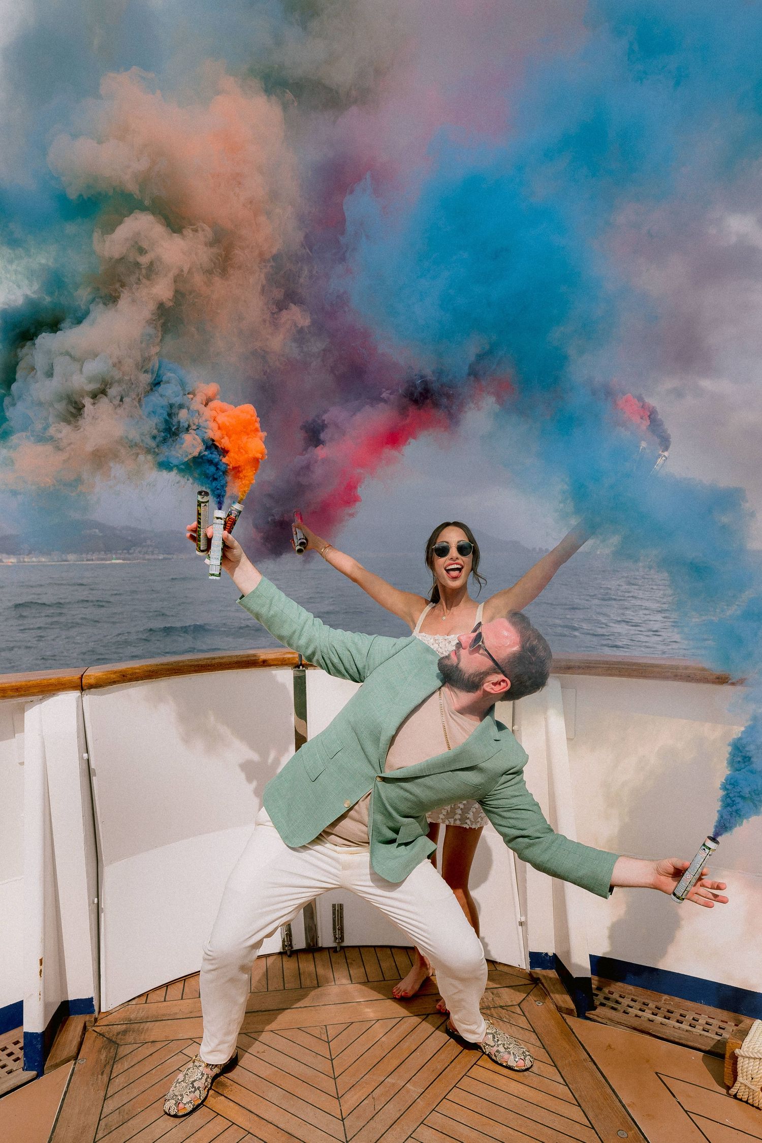 Two people celebrate on a boat deck with colorful smoke bombs creating vibrant clouds of blue, orange and pink in the sky.