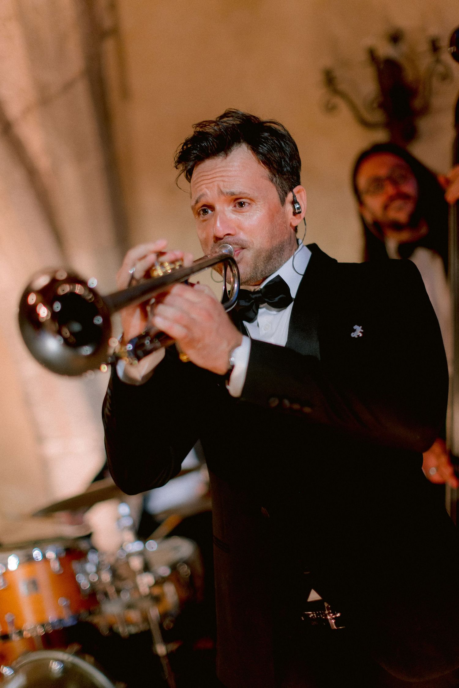 A trumpet player in a tuxedo performs at an elegant event with drums visible in the background.