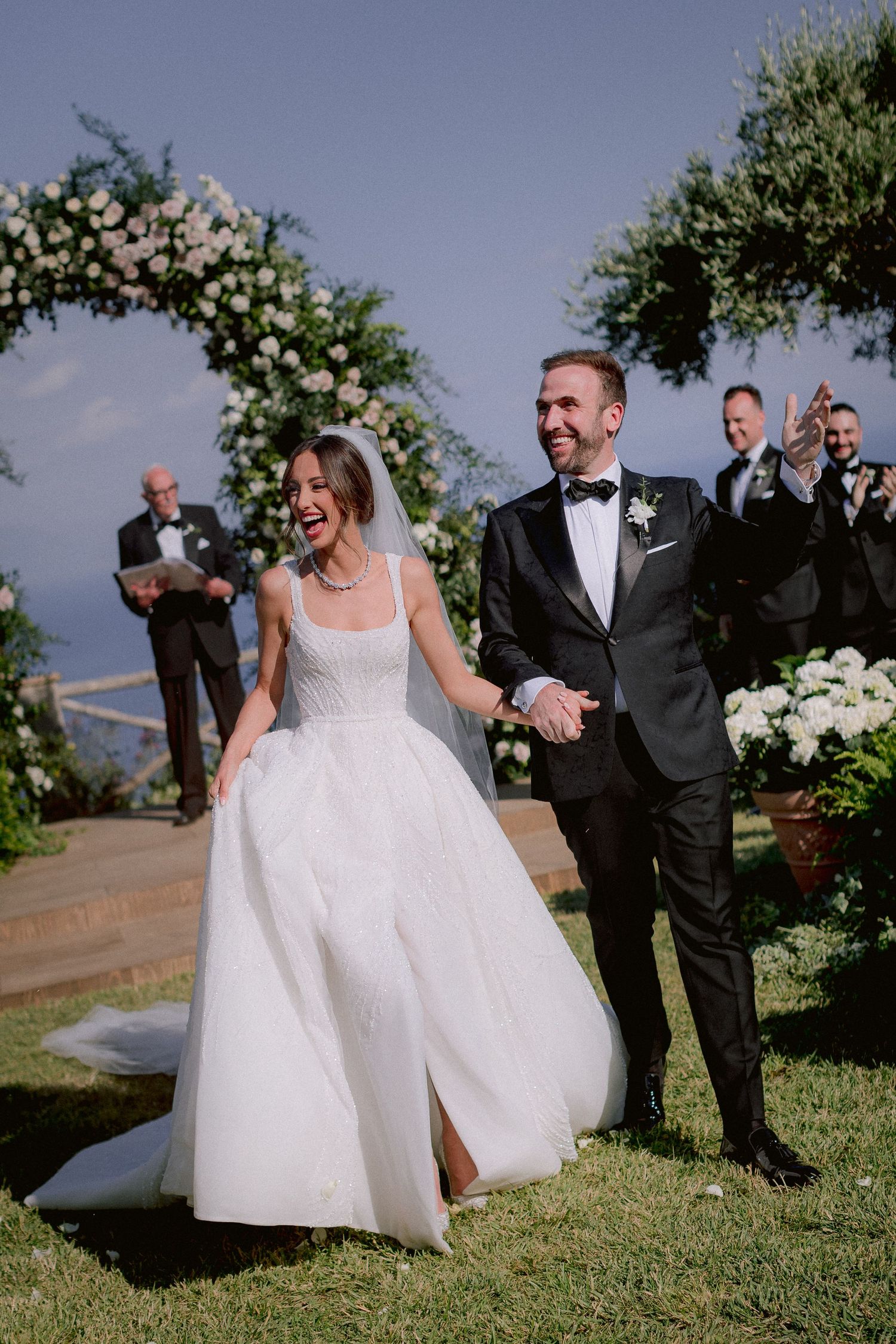 A wedding couple celebrates their ceremony under a floral arch at an outdoor venue with scenic views.