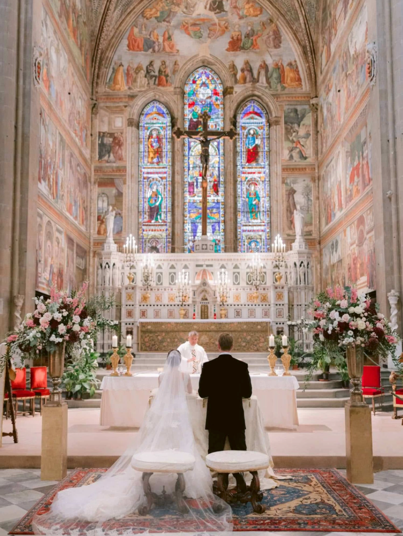 Bride and groom stand before ornate altar in historic church with stunning stained glass windows and elaborate frescoes.