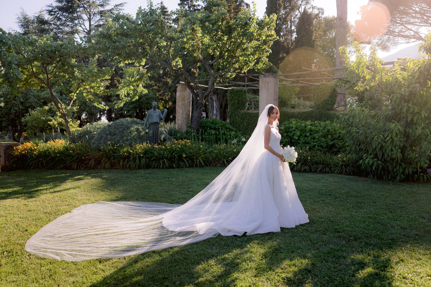 A flowing white wedding gown with long train sparkles in golden sunlight on a manicured garden lawn.