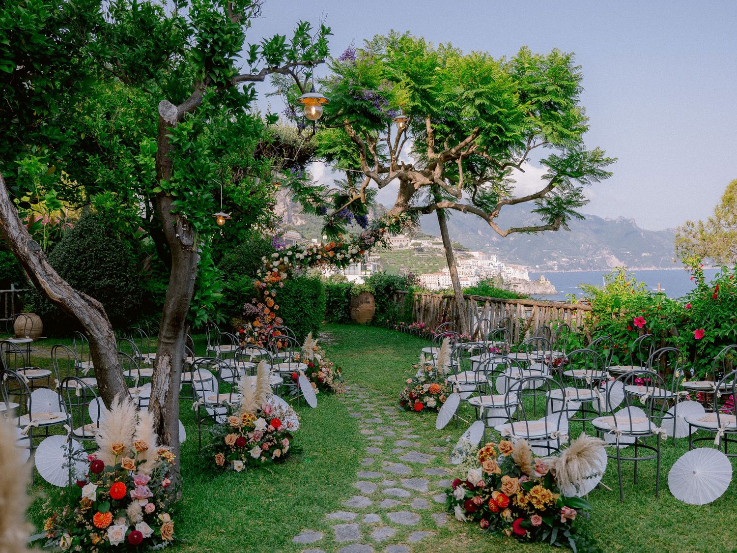 Outdoor wedding ceremony setup overlooking the coast with white chairs, floral arrangements and hanging lanterns in Italy.