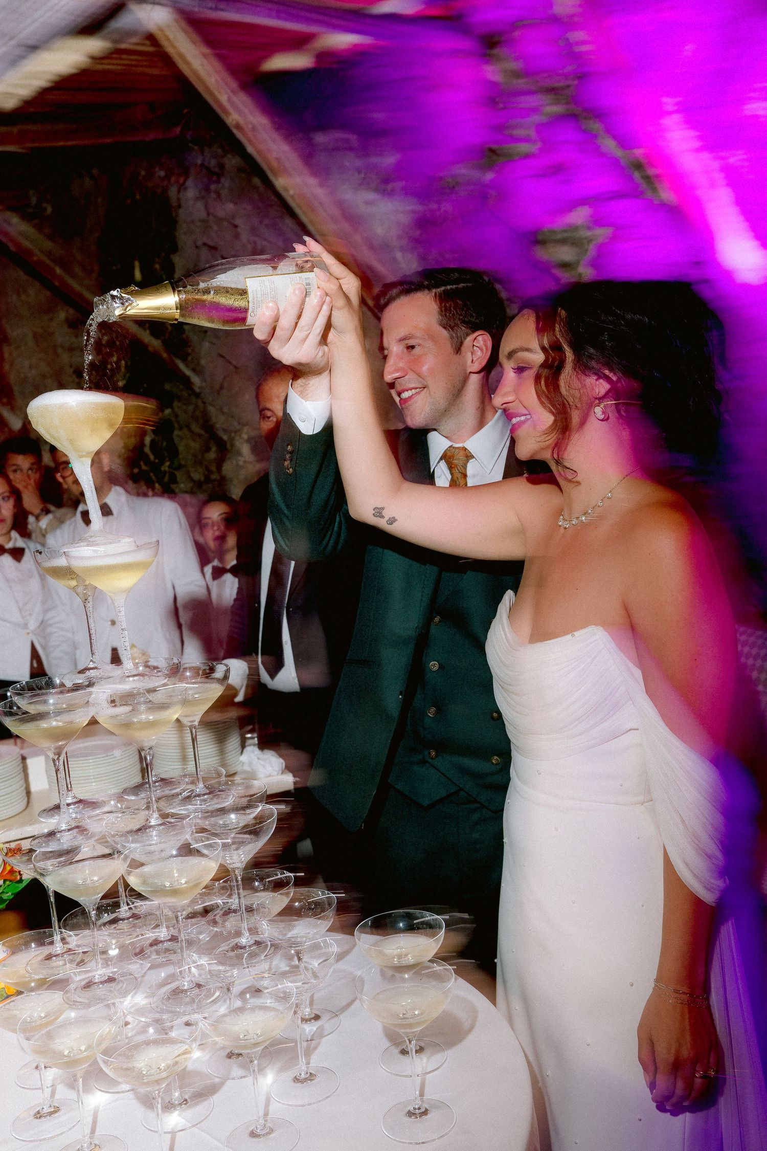 A couple pours champagne into a tower of cocktail glasses at an elegant wedding reception with purple lighting.