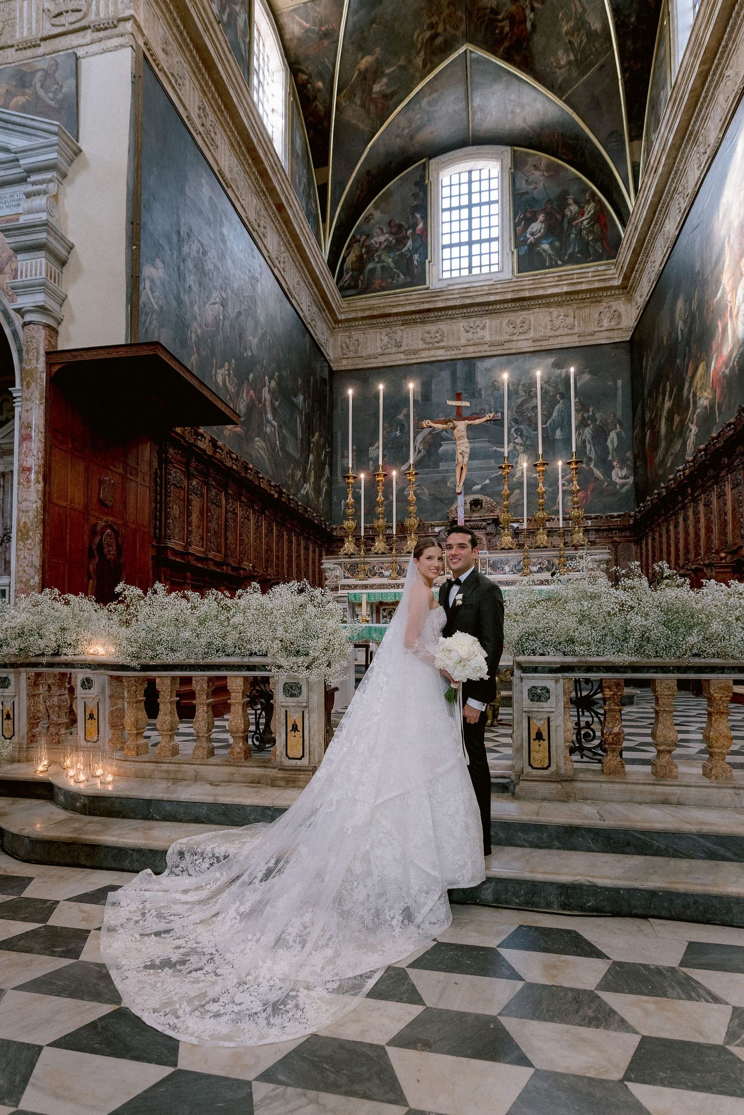 A wedding couple stands in a grand cathedral with ornate architecture, candles and baby's breath flowers surrounding them.