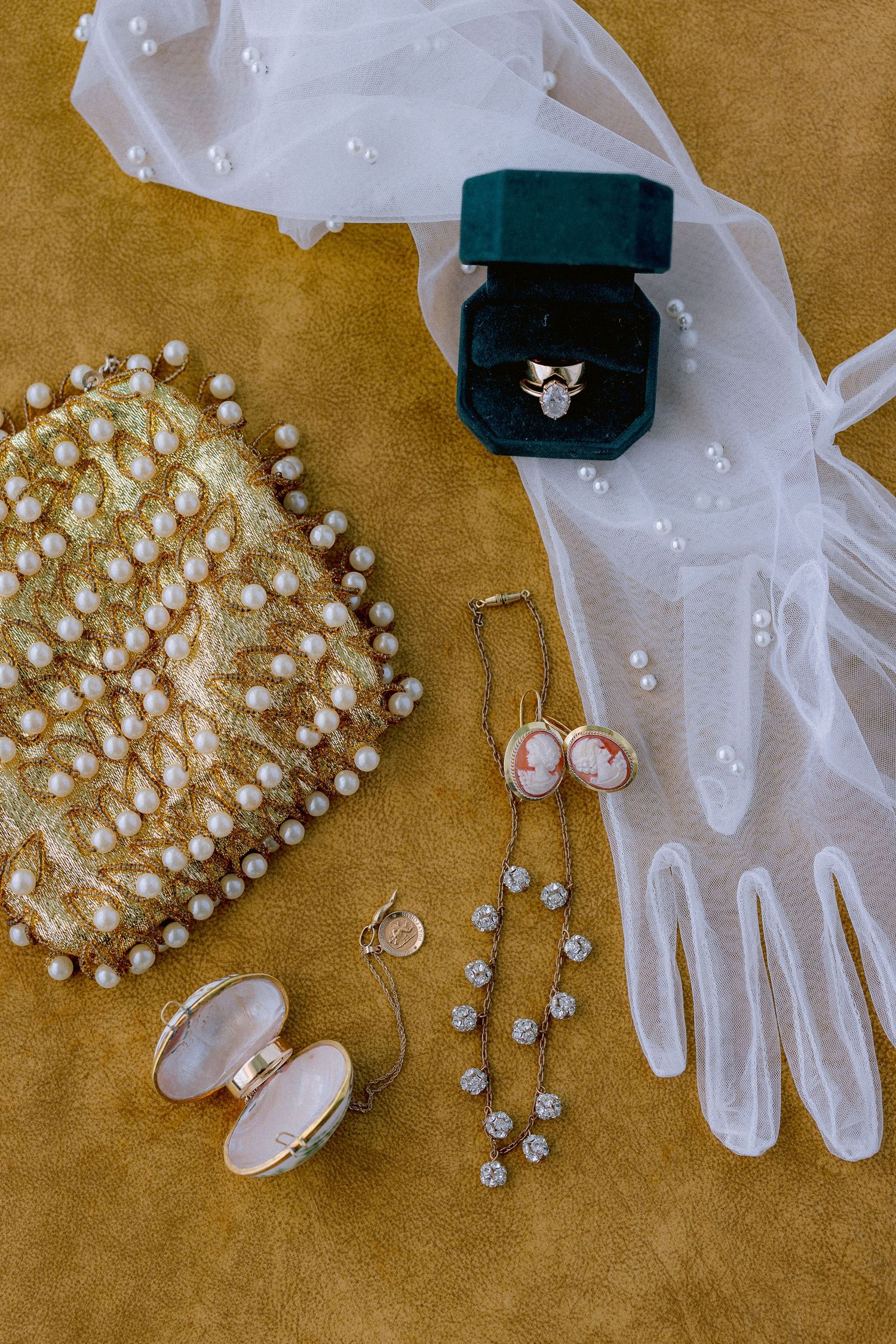 Elegant wedding accessories including pearl necklace, lace gloves, diamond ring box, and delicate jewelry on gold fabric.
