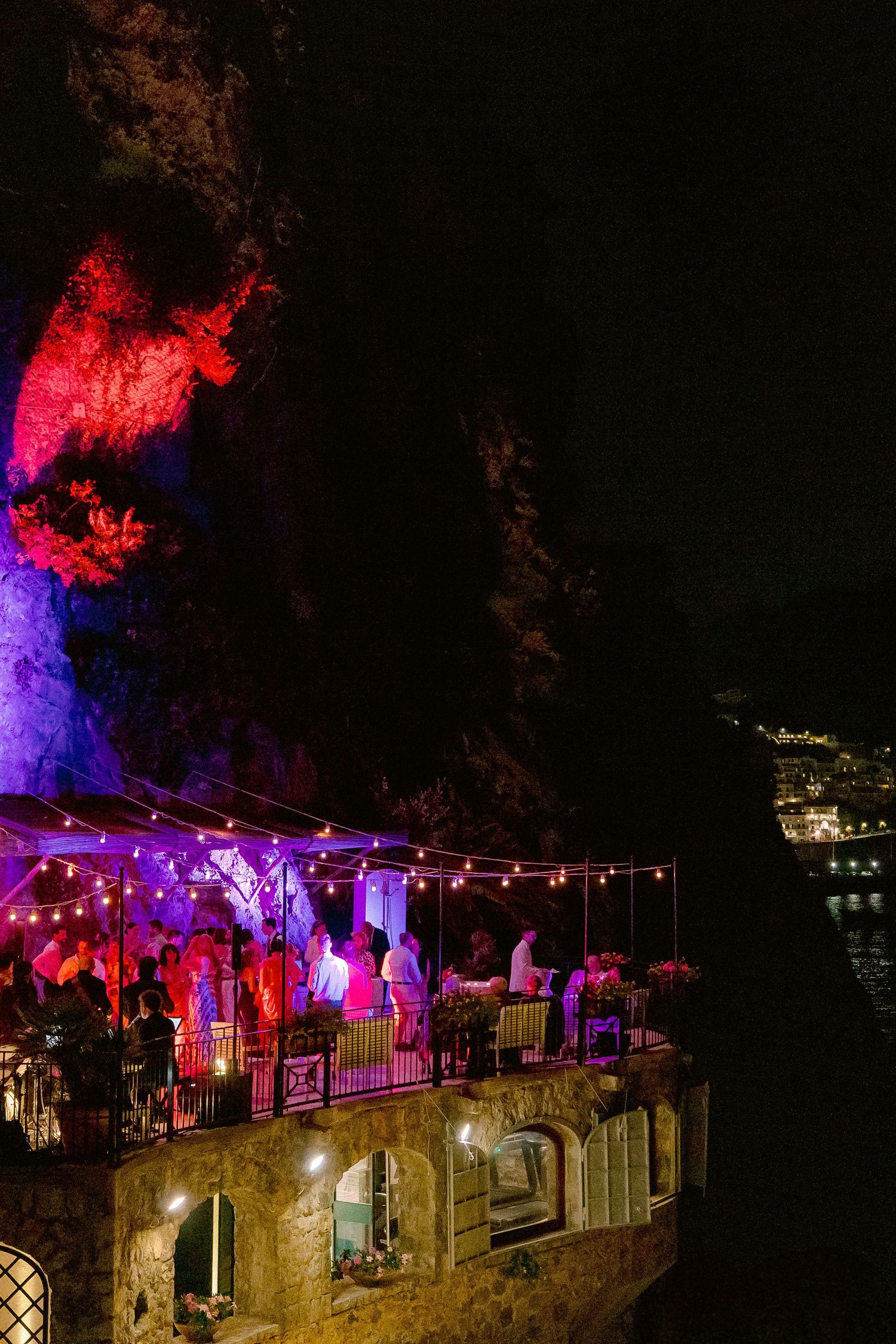 Nighttime party scene with trees illuminated in red and purple lights on a stone terrace or balcony.