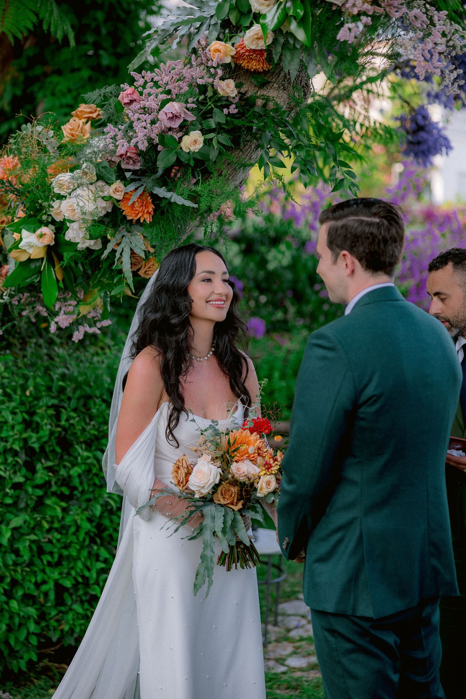 A romantic outdoor wedding ceremony takes place under a floral arch with orange and purple blooms in a lush garden setting.