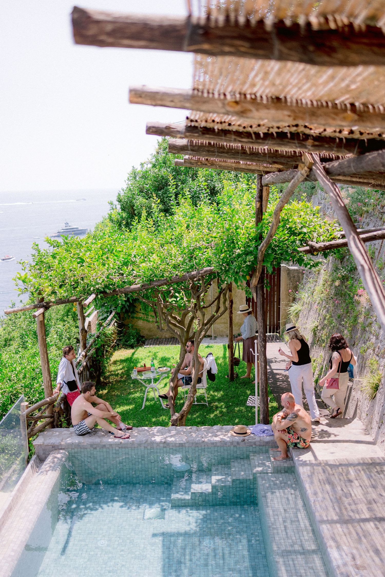 Rustic pool terrace with wood pergola and climbing vines overlooking scenic coastal views on a summer day.