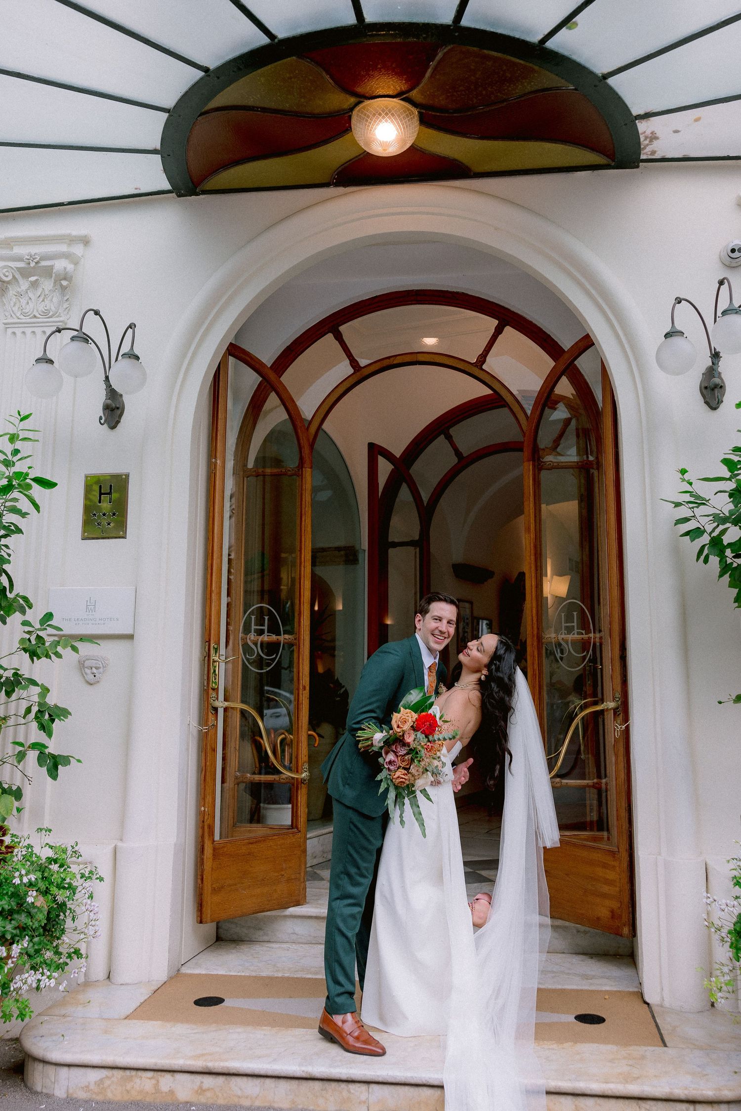 A couple stands in an elegant arched doorway with art deco details, surrounded by greenery and wall sconces.
