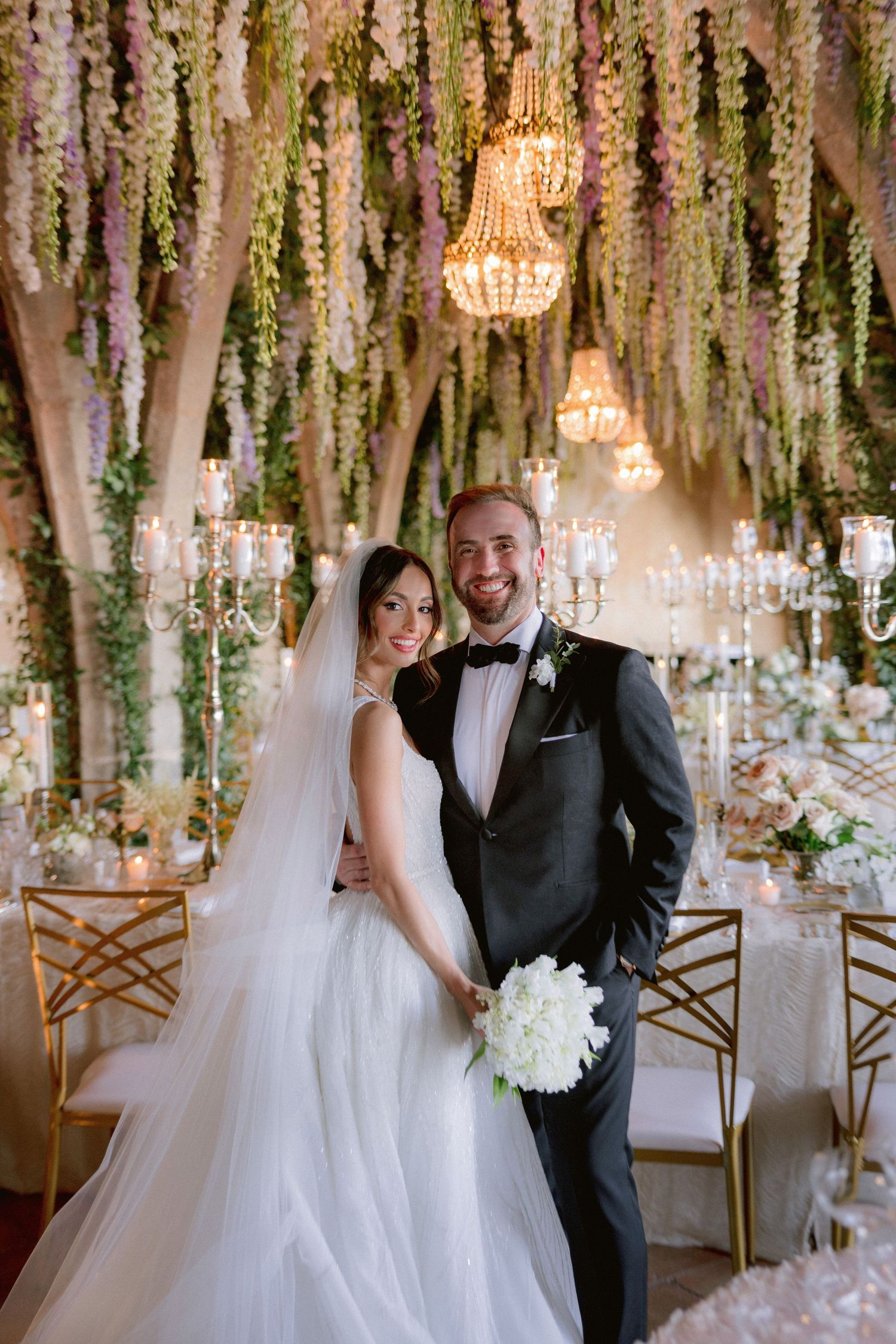 Bride and groom pose elegantly under floral ceiling with chandeliers in a beautifully decorated wedding reception venue.